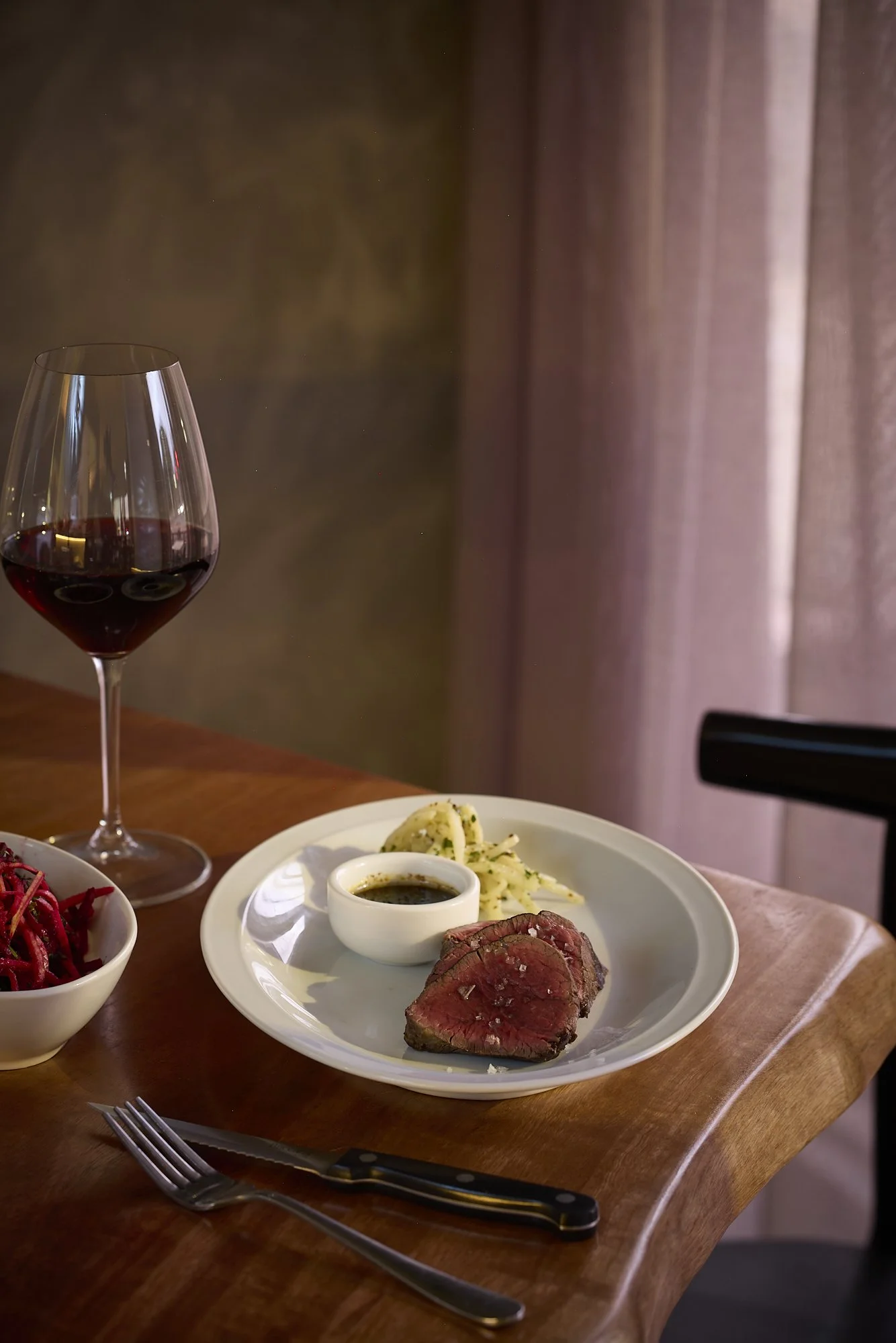 A dinner plate with slices of medium-rare steak, mashed potatoes, and a small bowl of gravy. A glass of red wine is beside the plate. A fork and knife are on the wooden table, with a curtain in the background.