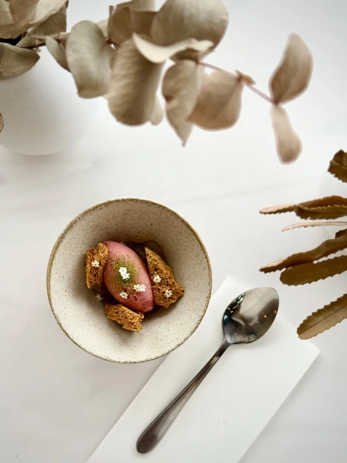 Dessert in a white bowl featuring a scoop of pink ice cream garnished with edible flowers, cookie pieces, and green sauce, placed on a white table with a silver spoon on a white napkin, with dried flowers in the background.