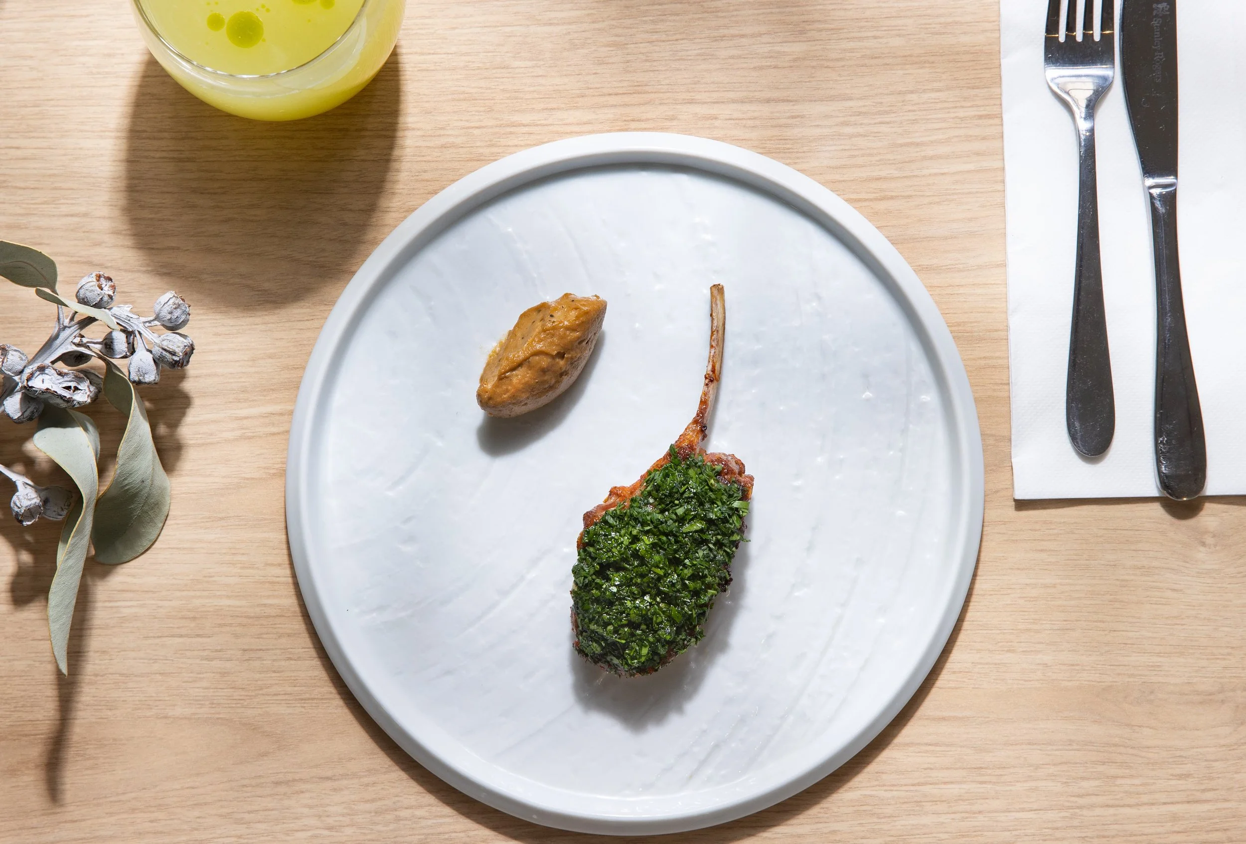 Plate with a lamb chop covered in herbs and a small dollop of sauce or puree, on a wooden table with a glass of yellow beverage, utensils on a napkin, and a sprig of greenery with berries nearby.