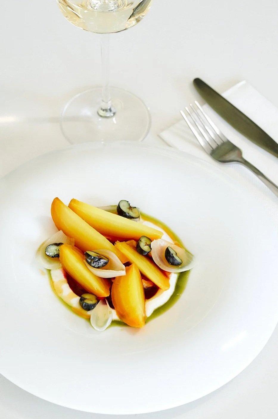 A white plate with a gourmet fruit salad featuring sliced peaches, melon, blueberries, and possibly some onion slices, garnished with a drizzle of green sauce, on a white table with a fork, knife, white napkin, and glass of white wine nearby.
