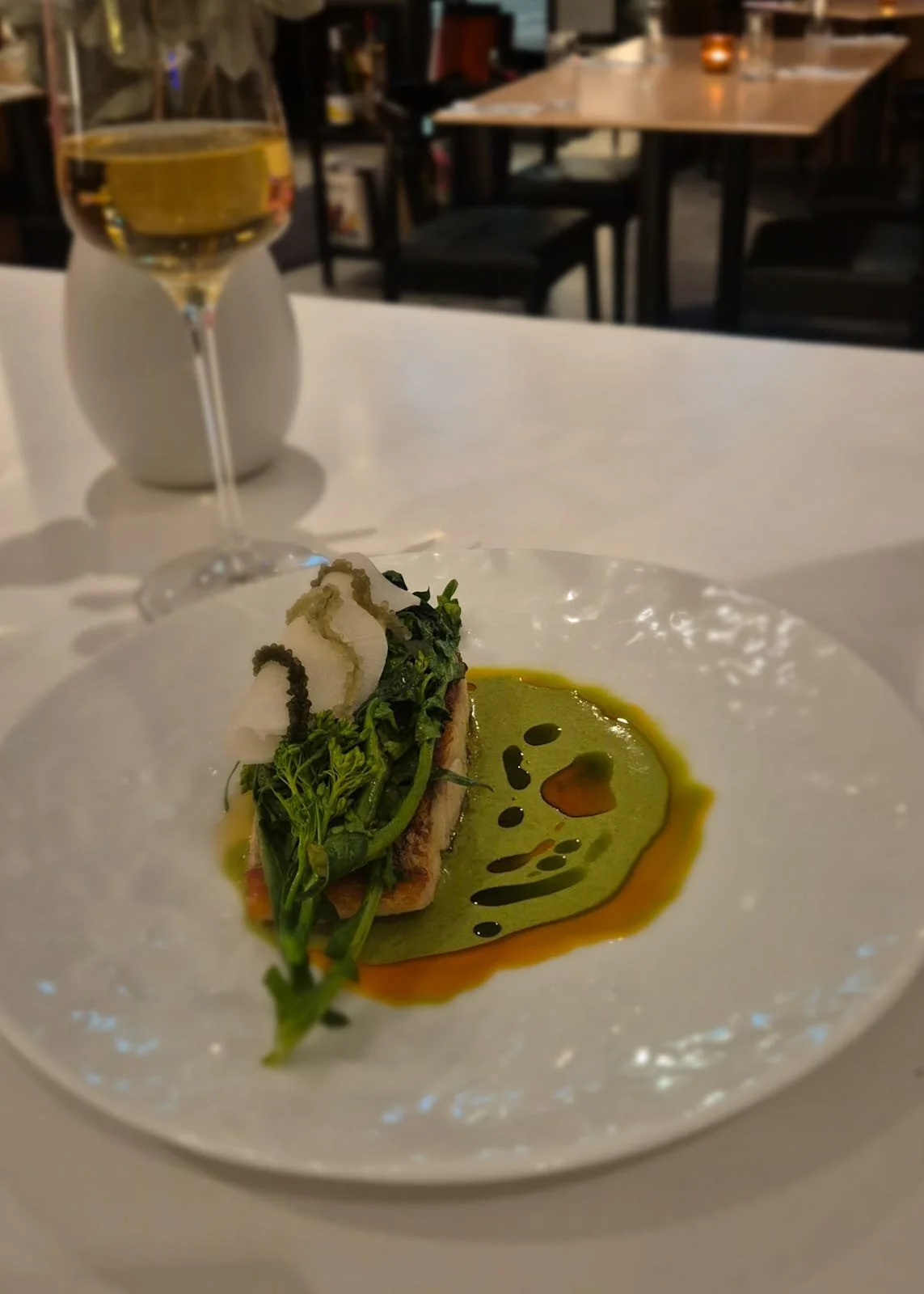 A gourmet dish with greens and a white decoration on a white plate, served with a green sauce and a brown sauce, in a restaurant with a glass of white wine in the background.