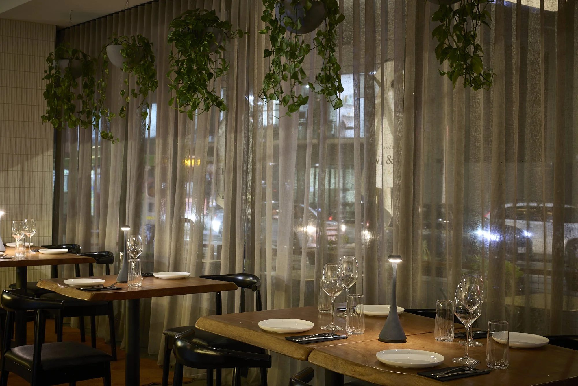 Interior of a restaurant with wooden tables set with wine glasses, plates, and silverware, near a large window with sheer curtains and hanging green plants.