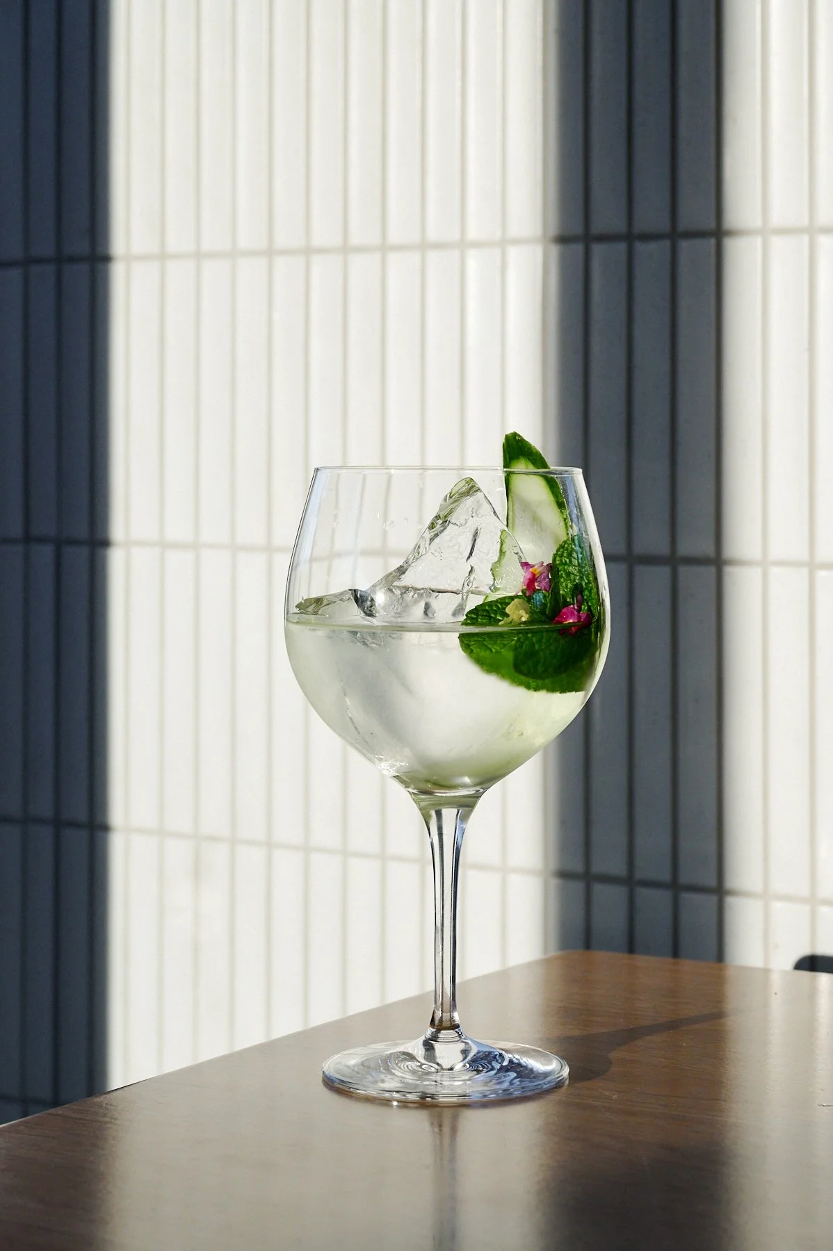 A glass of cocktail garnished with mint, cucumber, and edible flowers, with ice cubes inside, on a wooden table.