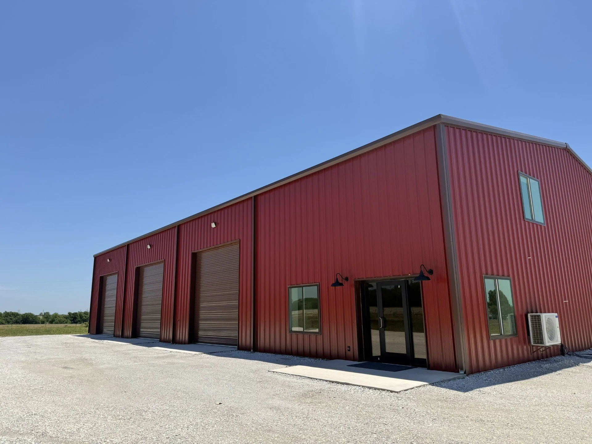Red metal building with large garage doors and small windows, set against a clear blue sky with some greenery in the background.