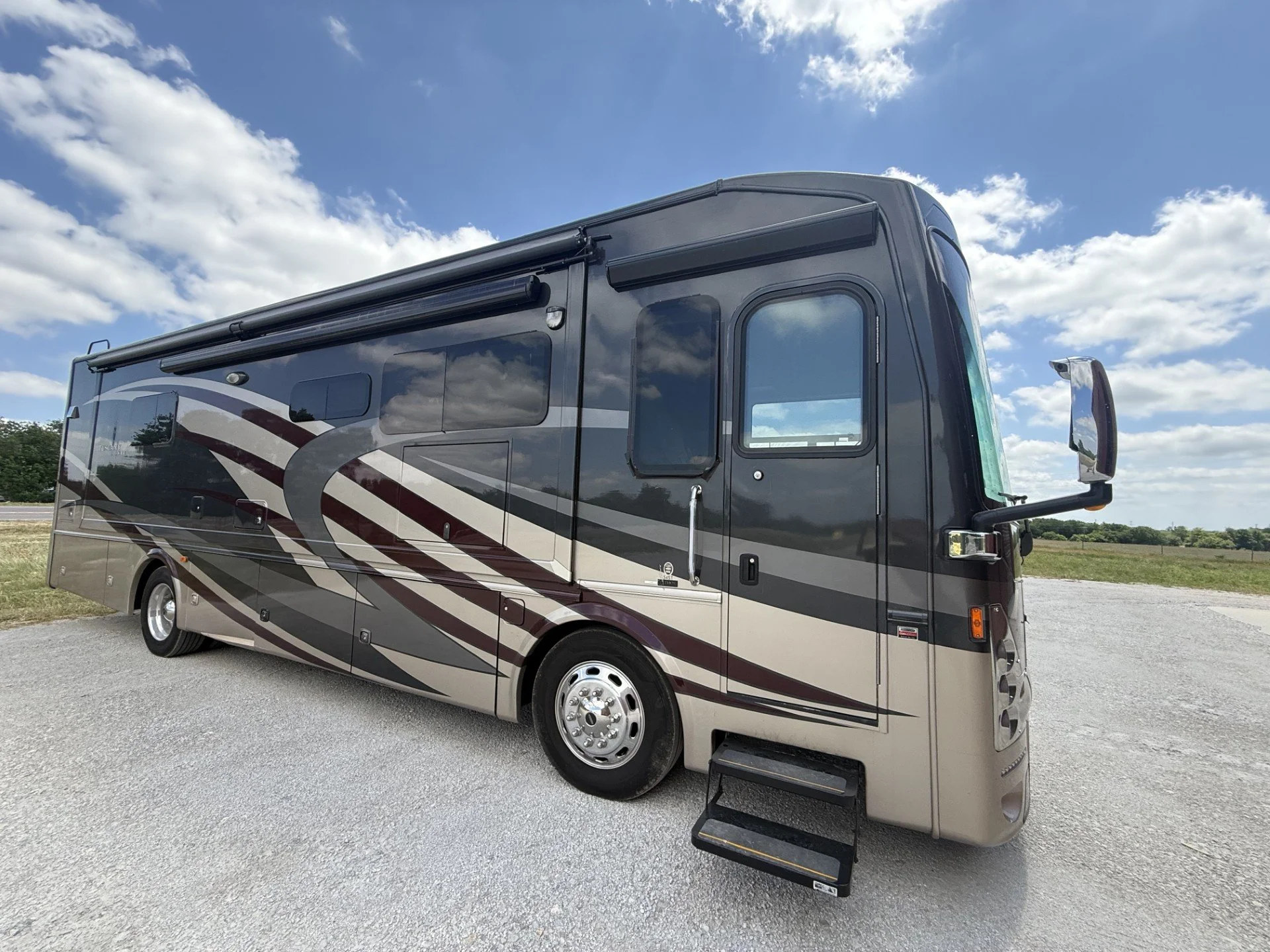 A large black, gray, and white motorhome with an extended step, parked on a gravel lot under a partly cloudy sky.