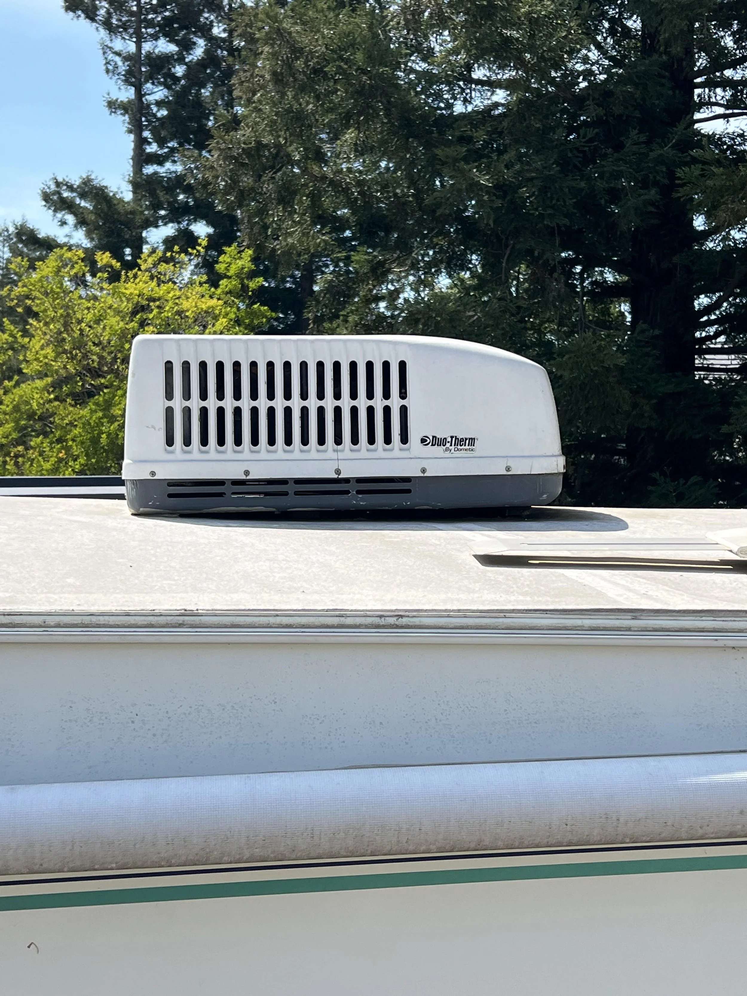 A white Duo-Therm air conditioner unit installed on the roof of a building with a background of green trees and a blue sky.