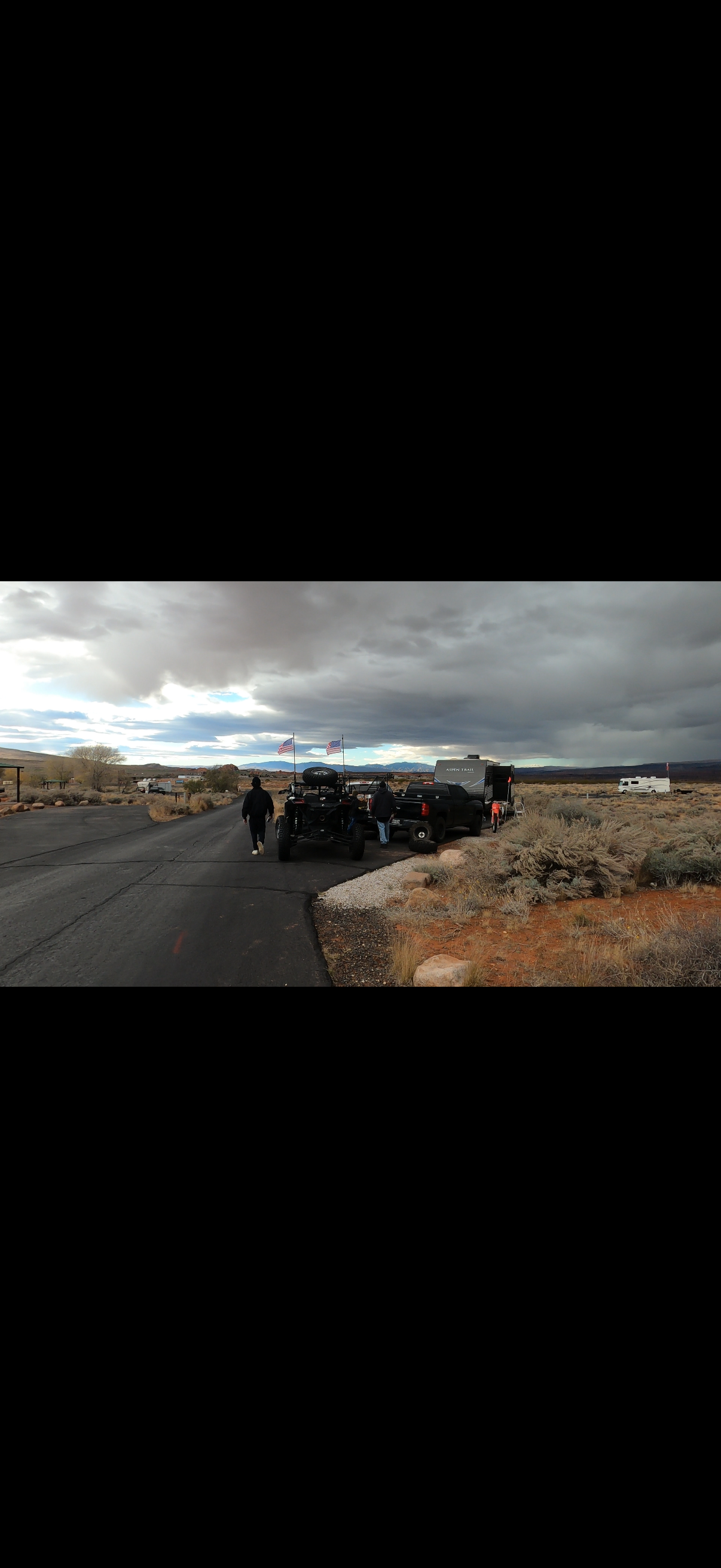 People standing near parked vehicles, including a black ATV, in a barren desert landscape under a cloudy sky with patches of blue.
