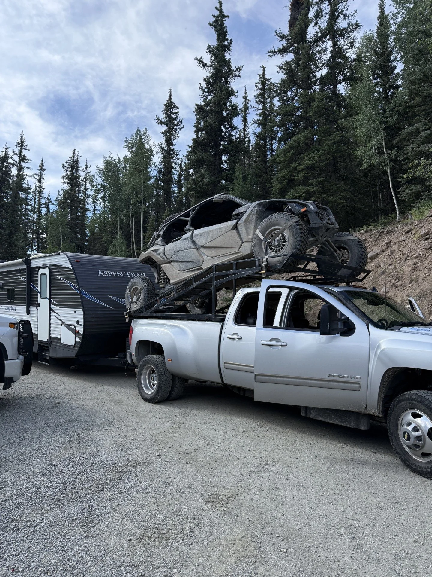 A silver pickup truck carrying a damaged off-road vehicle on its bed, parked next to a travel trailer against a backdrop of trees and a cloudy sky.