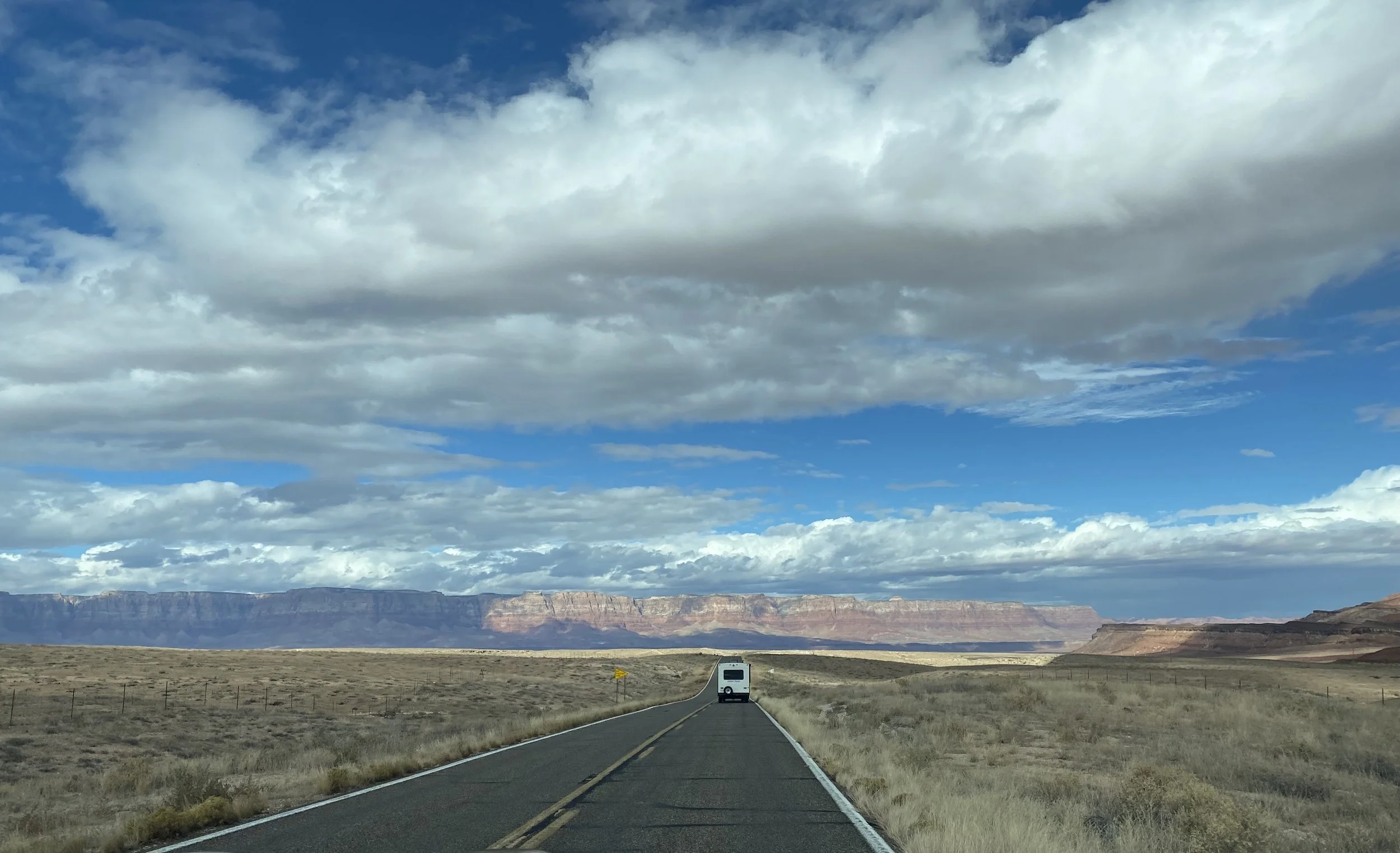A lone white vehicle driving down a straight two-lane road through a vast desert landscape with red rock mesas in the distance under a partly cloudy sky.