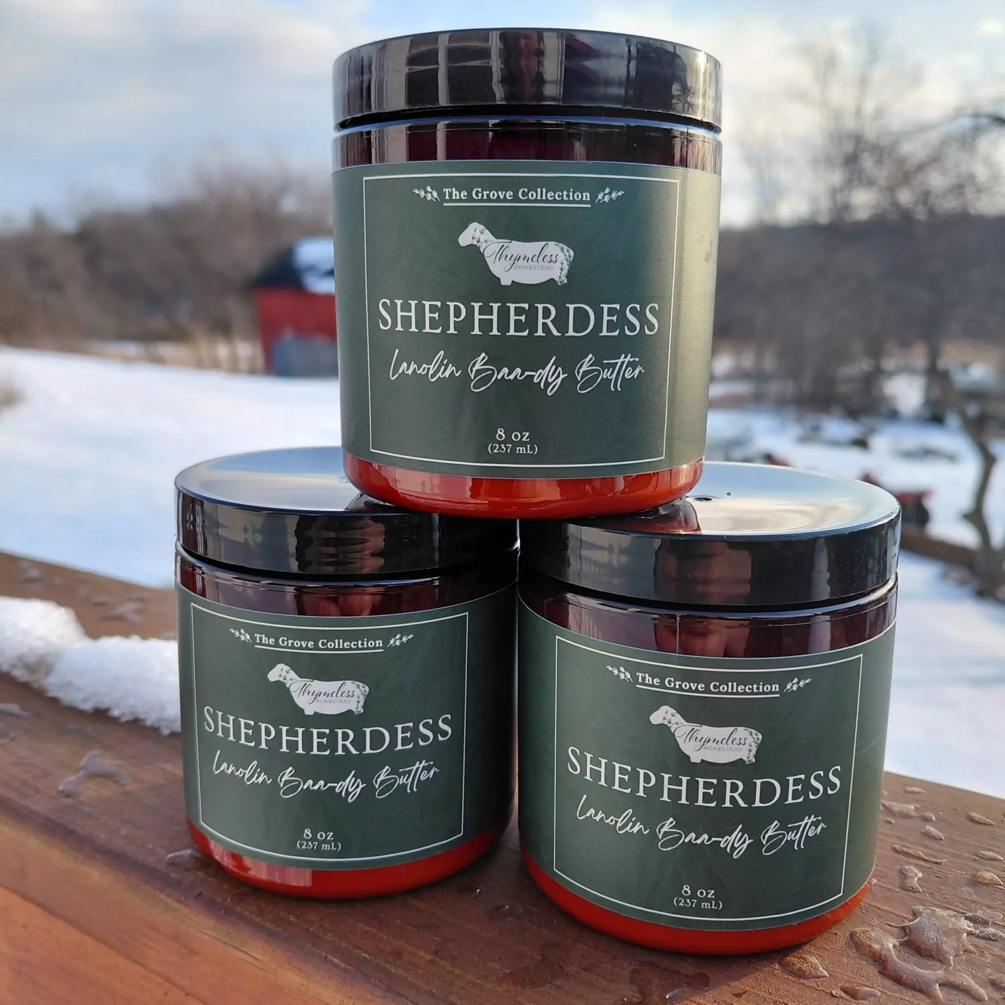 Three Shepherdess Lanolin Baa-dy Butters stacked in a pyramid, sitting on a wooden surface with snow and trees blurred in the background