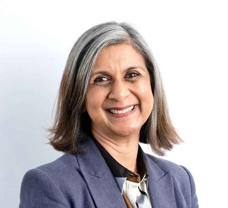 Portrait of a smiling middle-aged woman with shoulder-length gray hair, wearing a navy blazer and patterned blouse, against a plain white background.