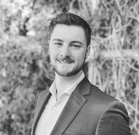 Young man with short dark hair, beard, wearing a suit and collared shirt, smiling outdoors with trees in background.