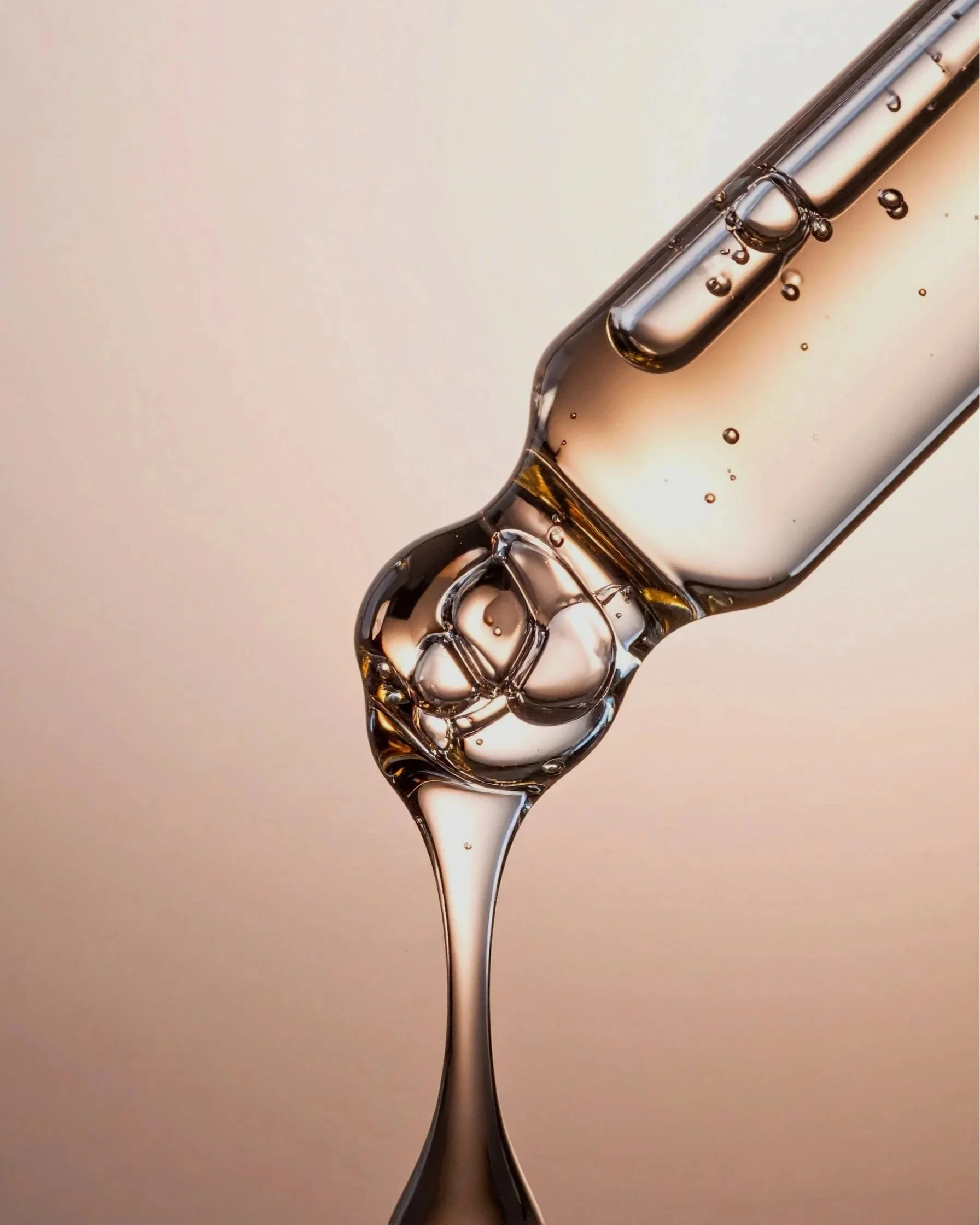 Close up image of a pipette dropper filled with clear thick serum. The background is a light pink colour