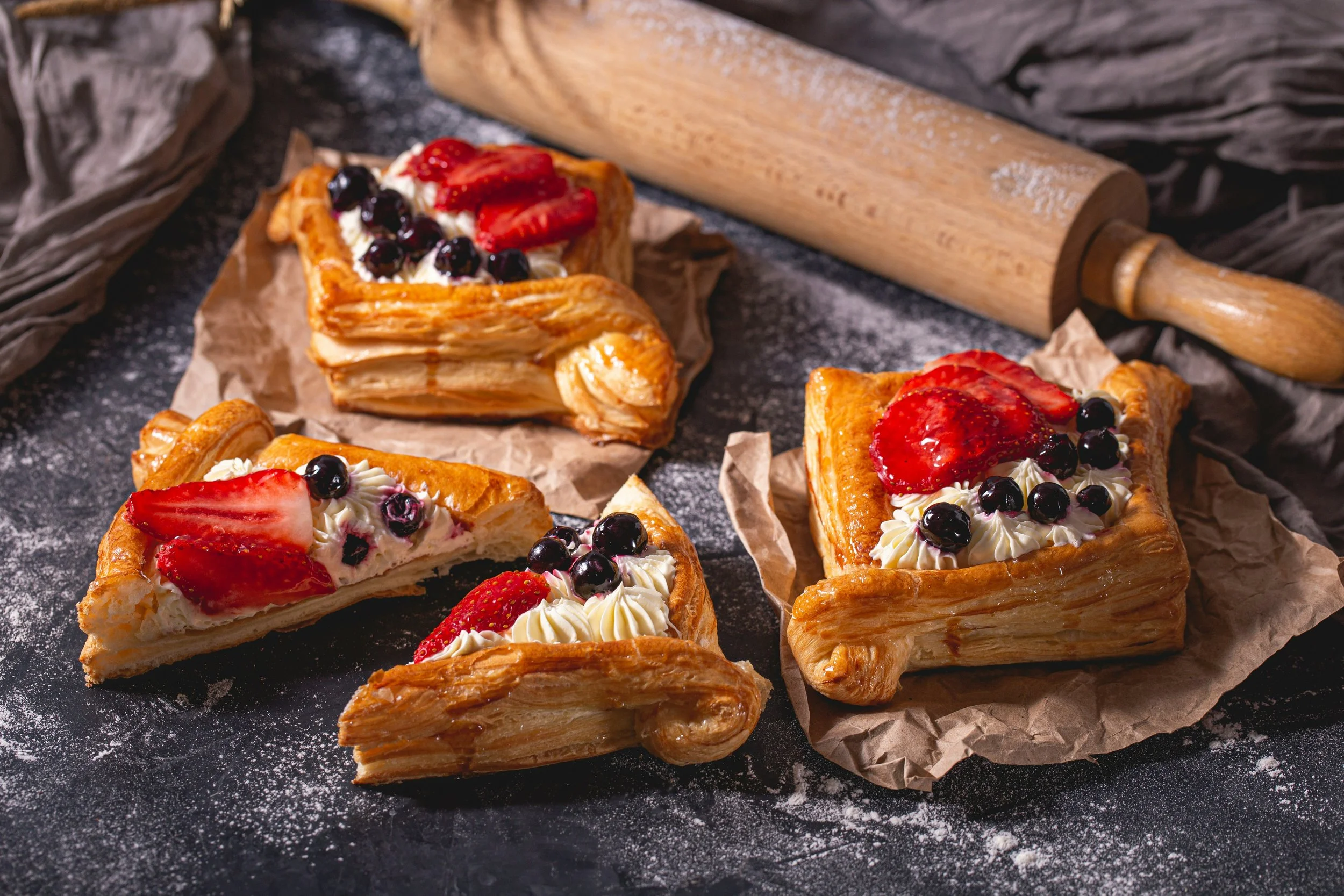 Three slices of fruit Danish pastry topped with strawberries, blueberries, and whipped cream on a black surface with flour, a rolling pin, and crumpled parchment paper.