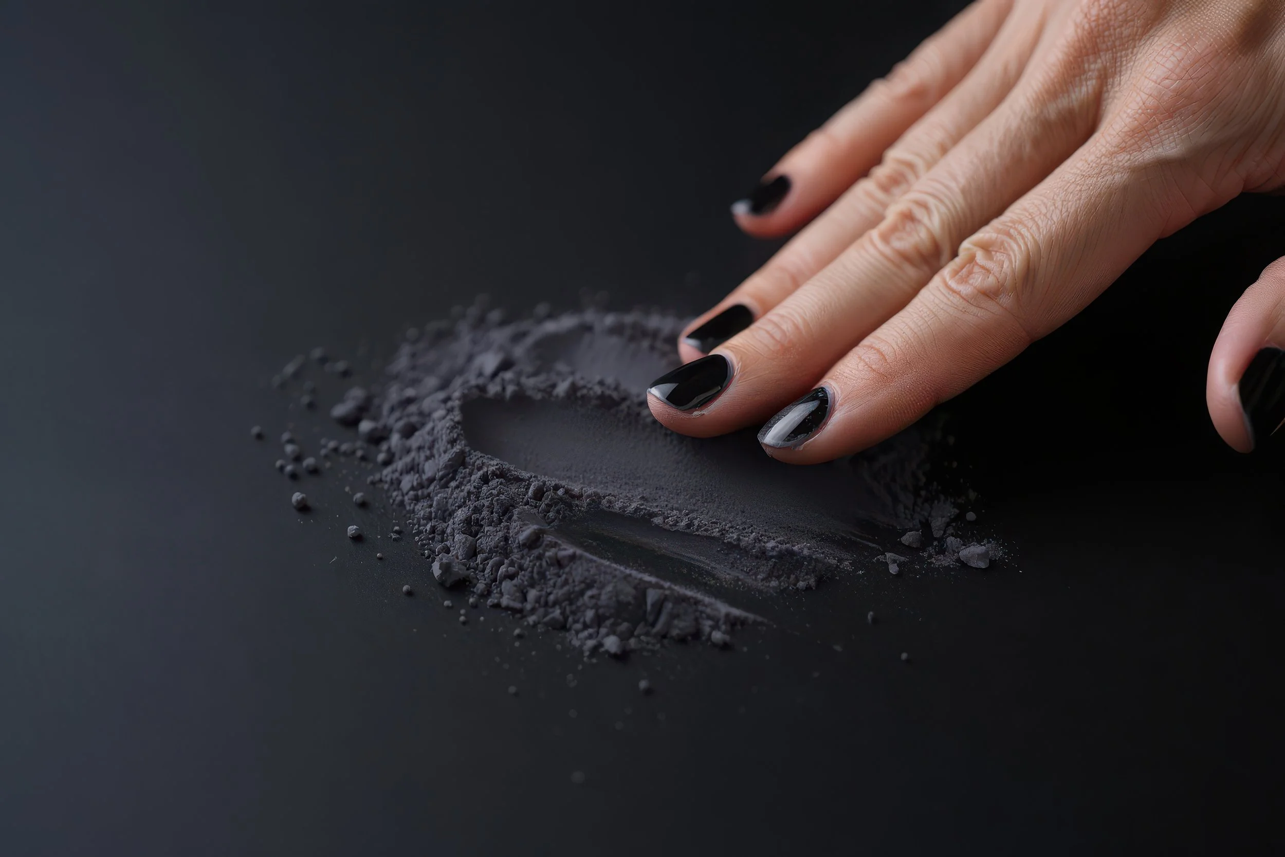 A hand with black nail polish presses onto a black surface, creating a smudge of black powder or pigment with surrounding scattered particles.