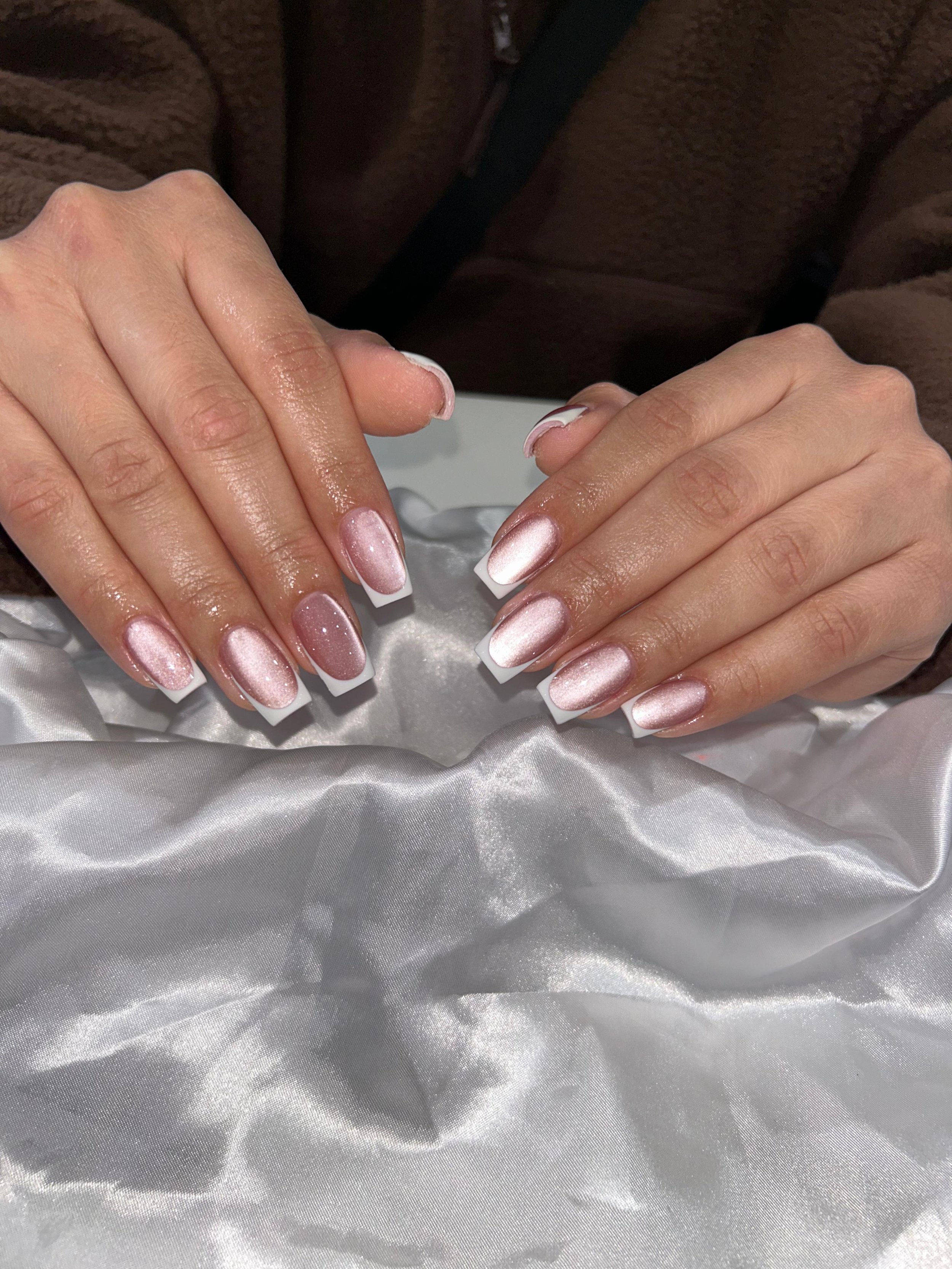 Close-up of hands with freshly manicured nails featuring metallic pink polish with white tips, resting on silky white fabric.