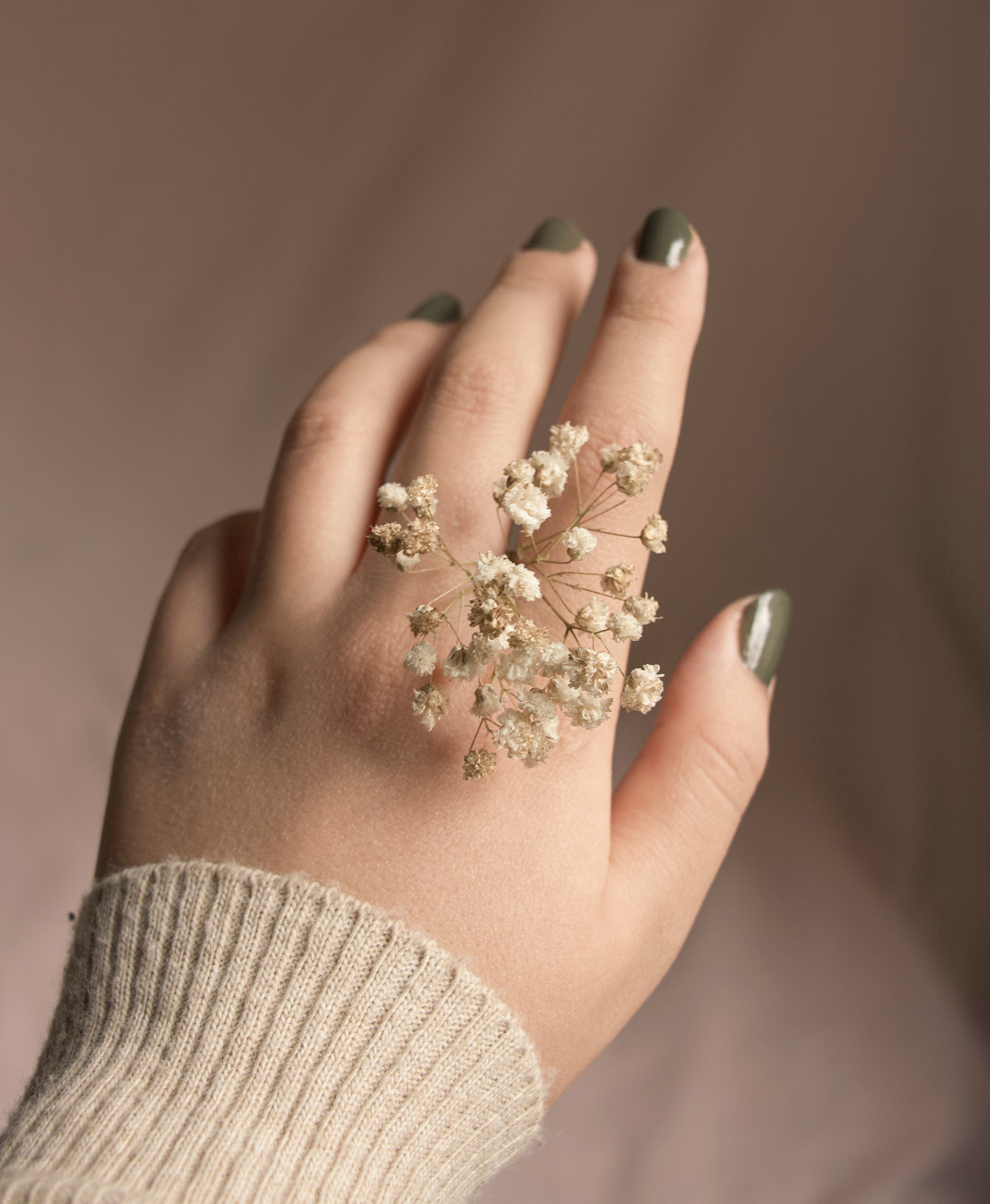 A hand with green nail polish and a beige knit sweater, with a small cluster of white dried flowers resting on the fingers.