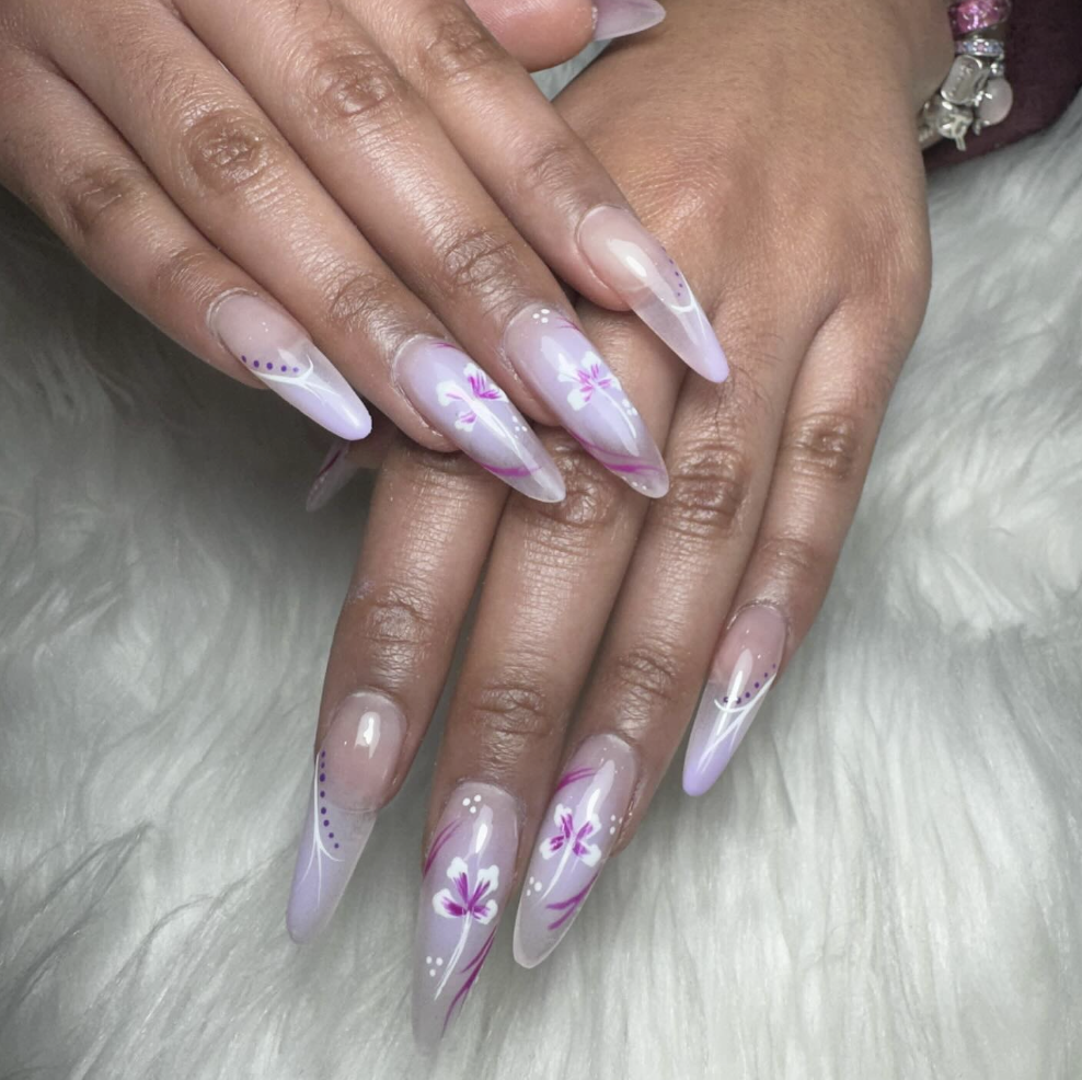 Close-up of hands with long, almond-shaped nails painted with light pink and purple floral nail art and white accents, resting on a furry surface.