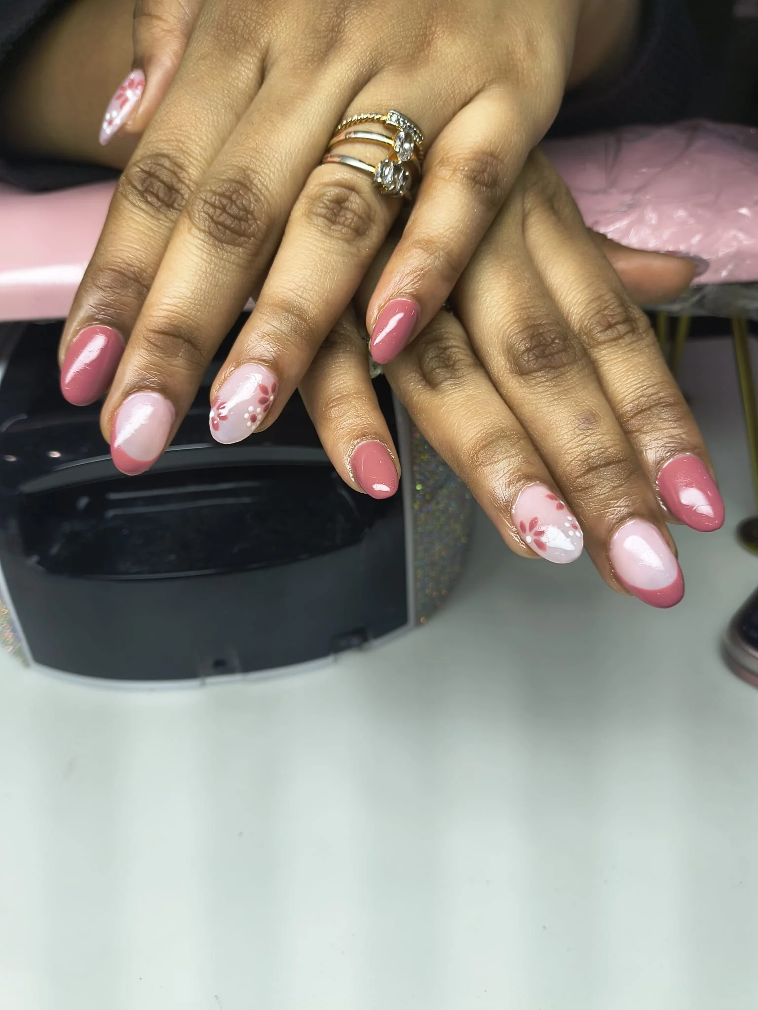 Hands with manicured nails resting on a black surface, displaying pink and white nail art with floral designs and jewelry rings on fingers.