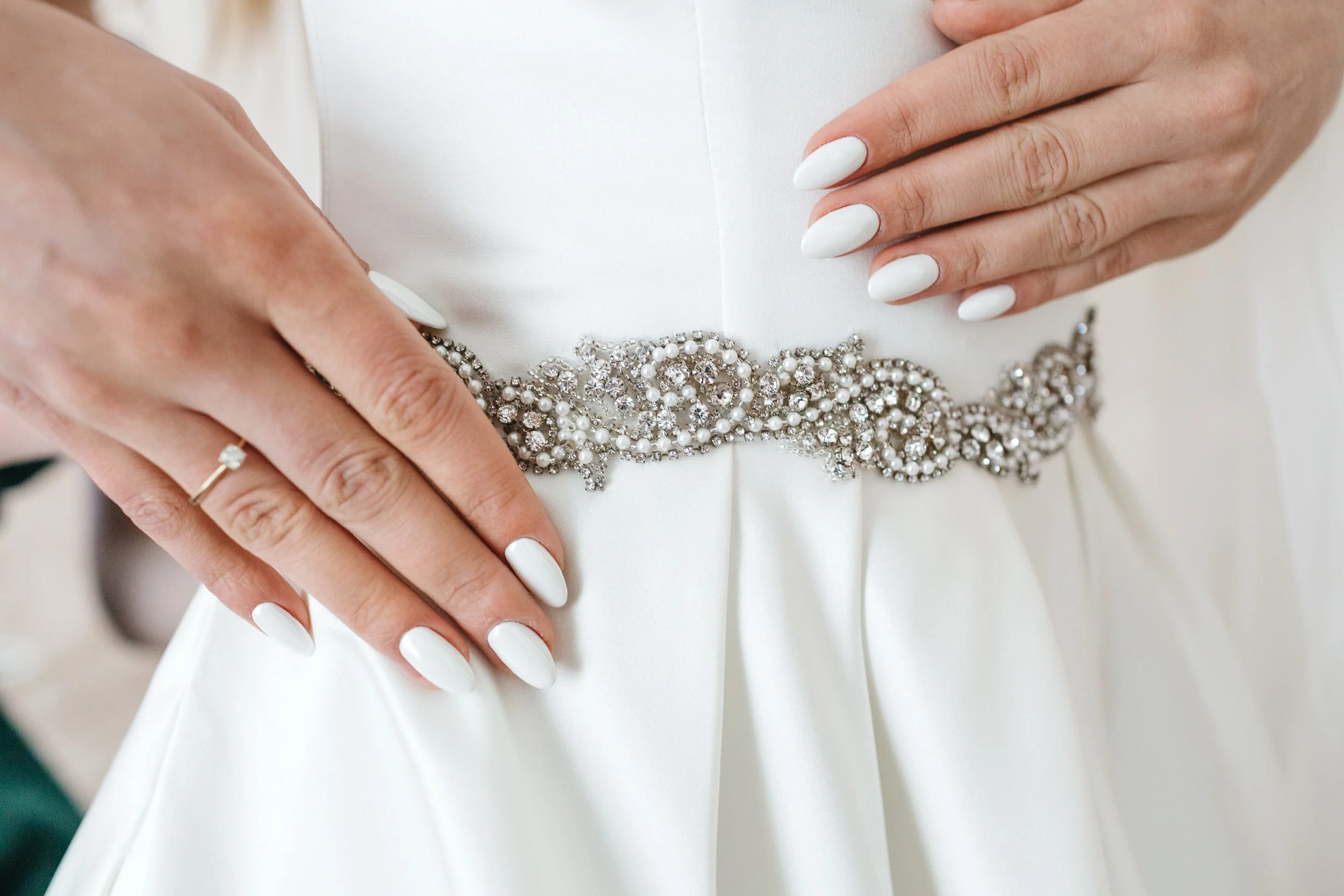 Hands of a bride with white nails and a ring, adjusting a bejeweled belt on a wedding dress.