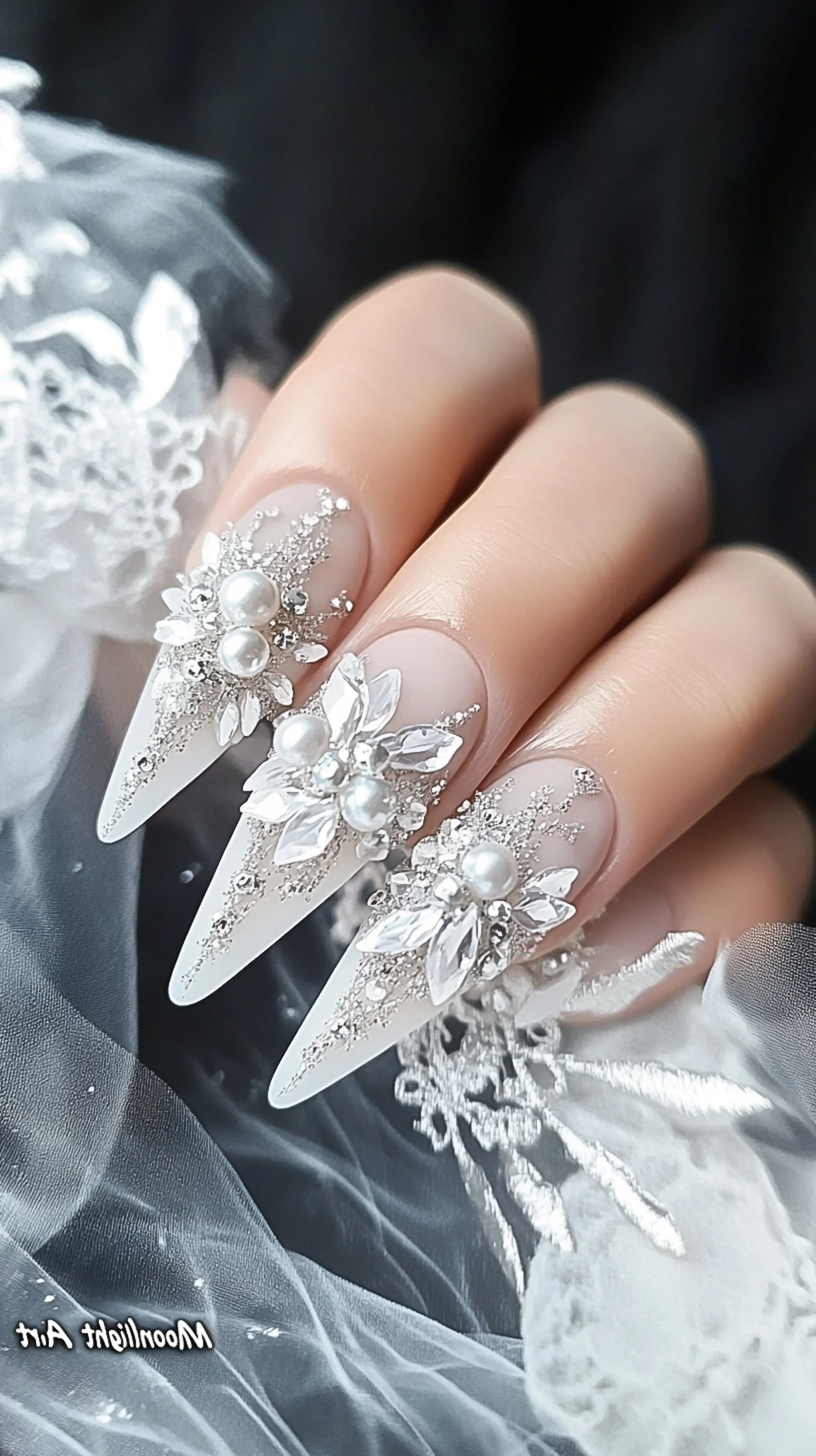 Close-up of elaborately decorated long white stiletto nails with rhinestones, pearls, and floral designs, held over a silver and white fabric background.