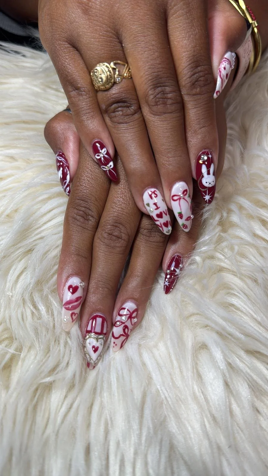 Close-up of a person's hand with festive Christmas-themed nail art, featuring red, white, and pink designs including hearts, bows, a bunny, and HOLIDAY words, resting on a fluffy white surface, wearing gold rings.