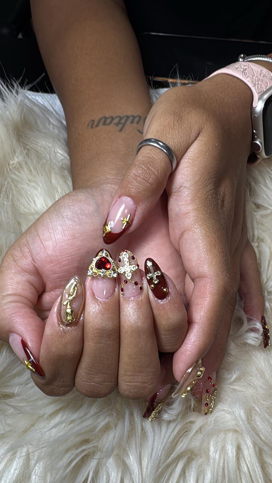 Close-up of two hands intertwined, showing manicured nails with red, gold, and clear designs, adorned with gold jewelry and rings, resting on a fluffy white surface.
