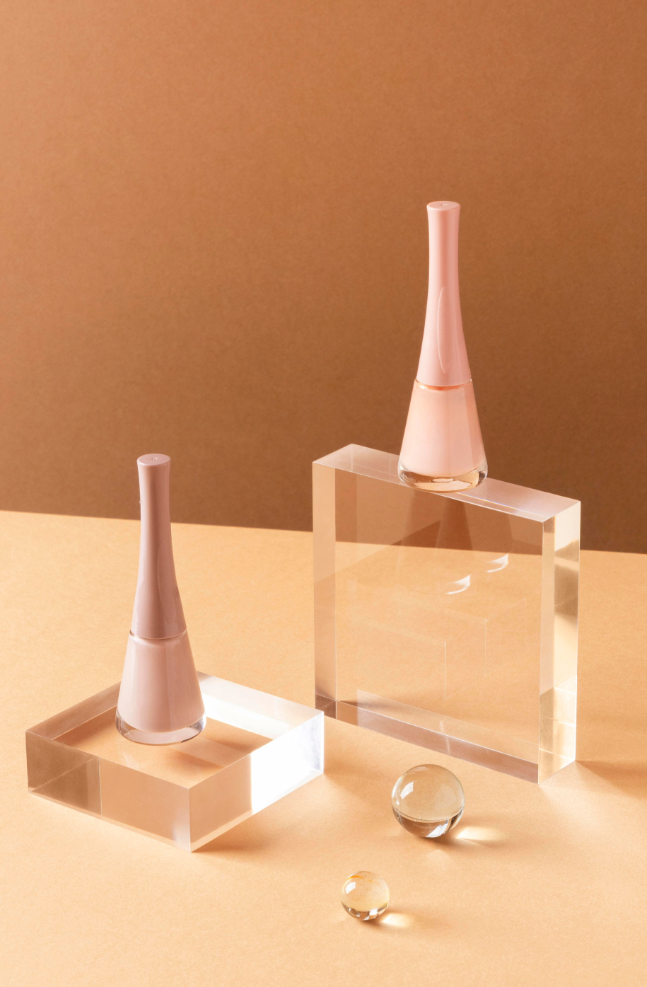 Two pink nail polish bottles on clear acrylic display blocks, with glass spheres on a peach-colored surface, against a peach background.