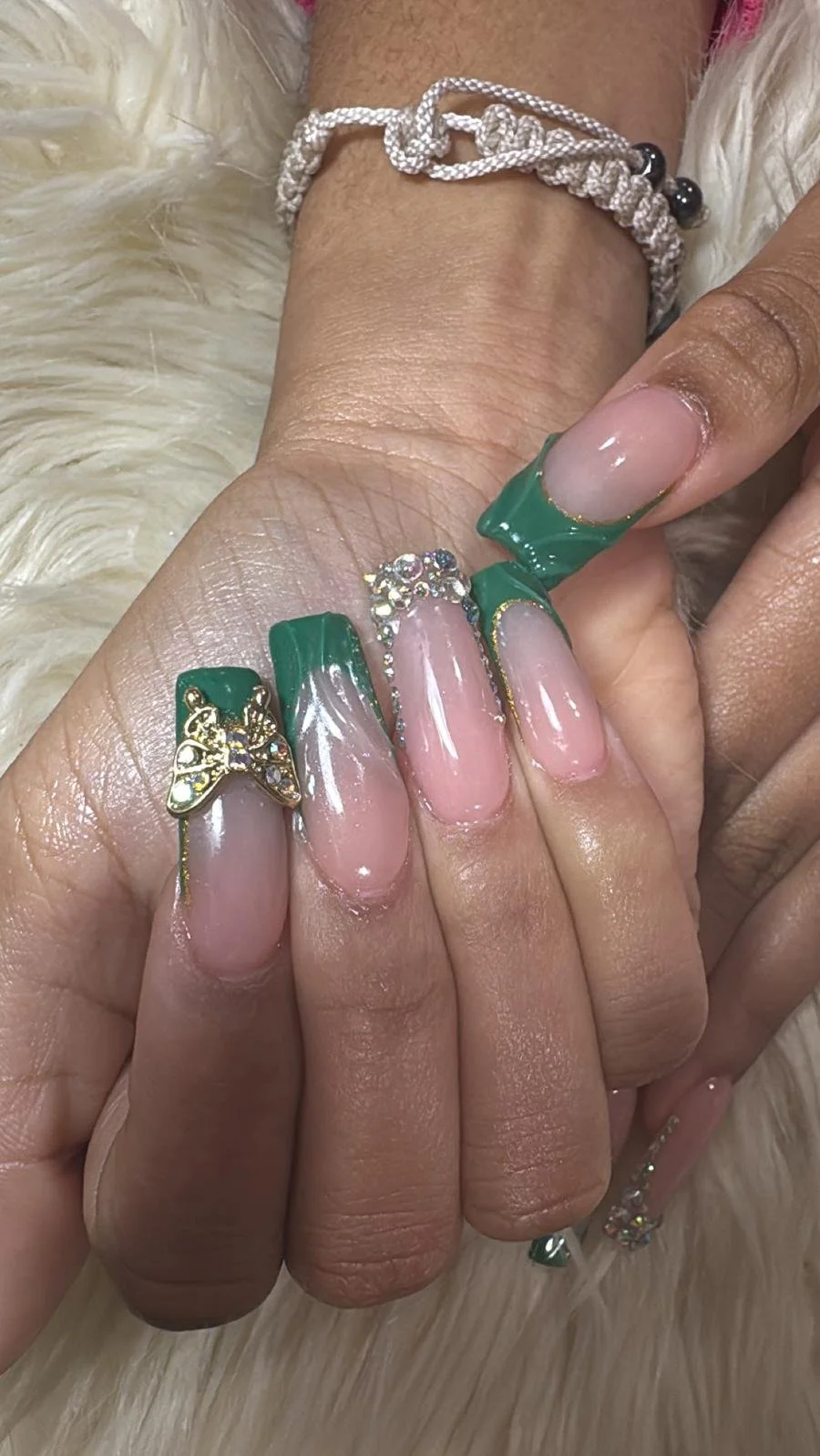 Close-up of a hand with long, decorated nails and jewelry, including a green french manicure with gold accents, rhinestones, and butterfly charms.