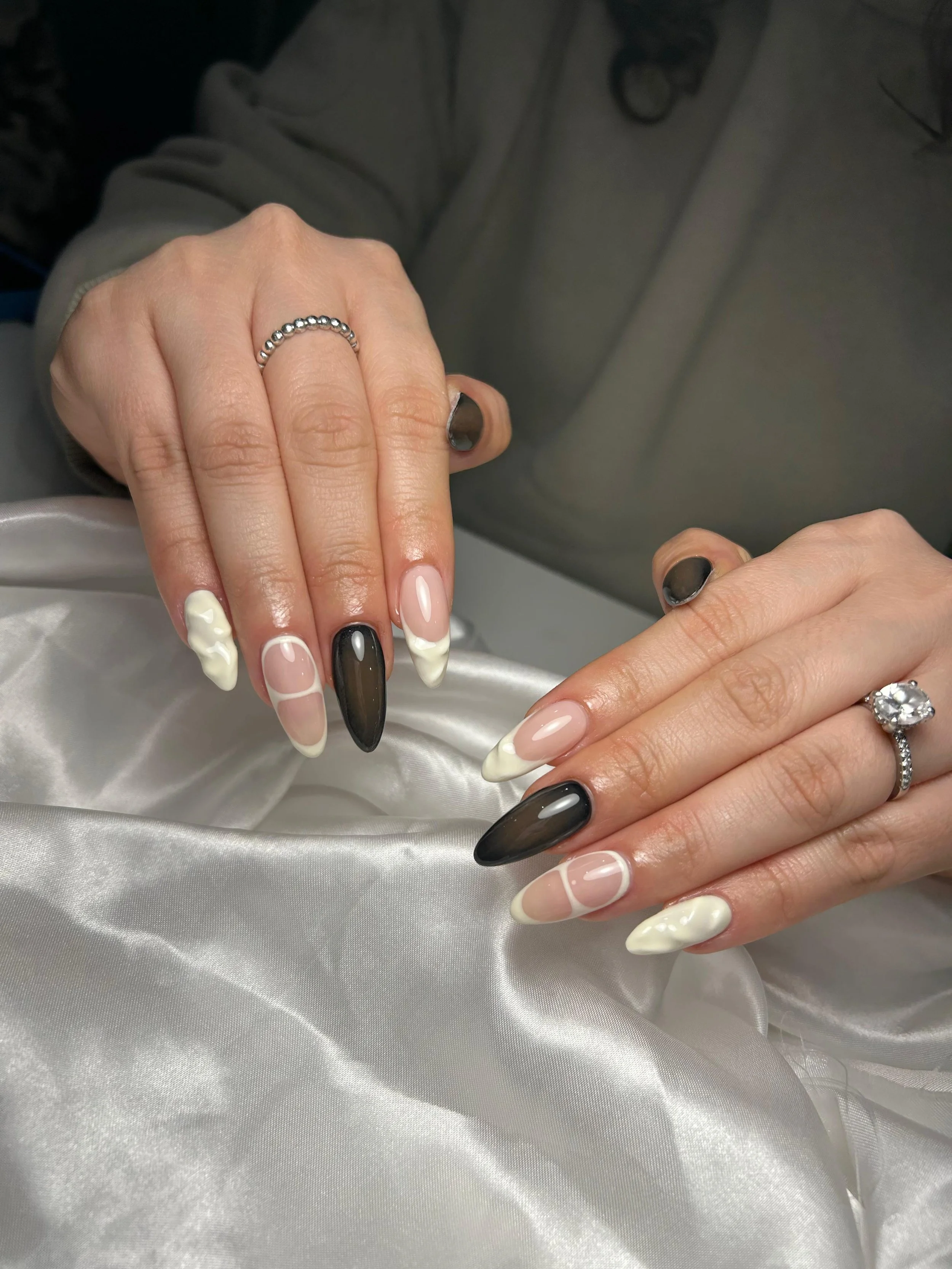Close-up of hands with manicured nails in black, white, and clear polish, resting on silky white fabric, adorned with rings.