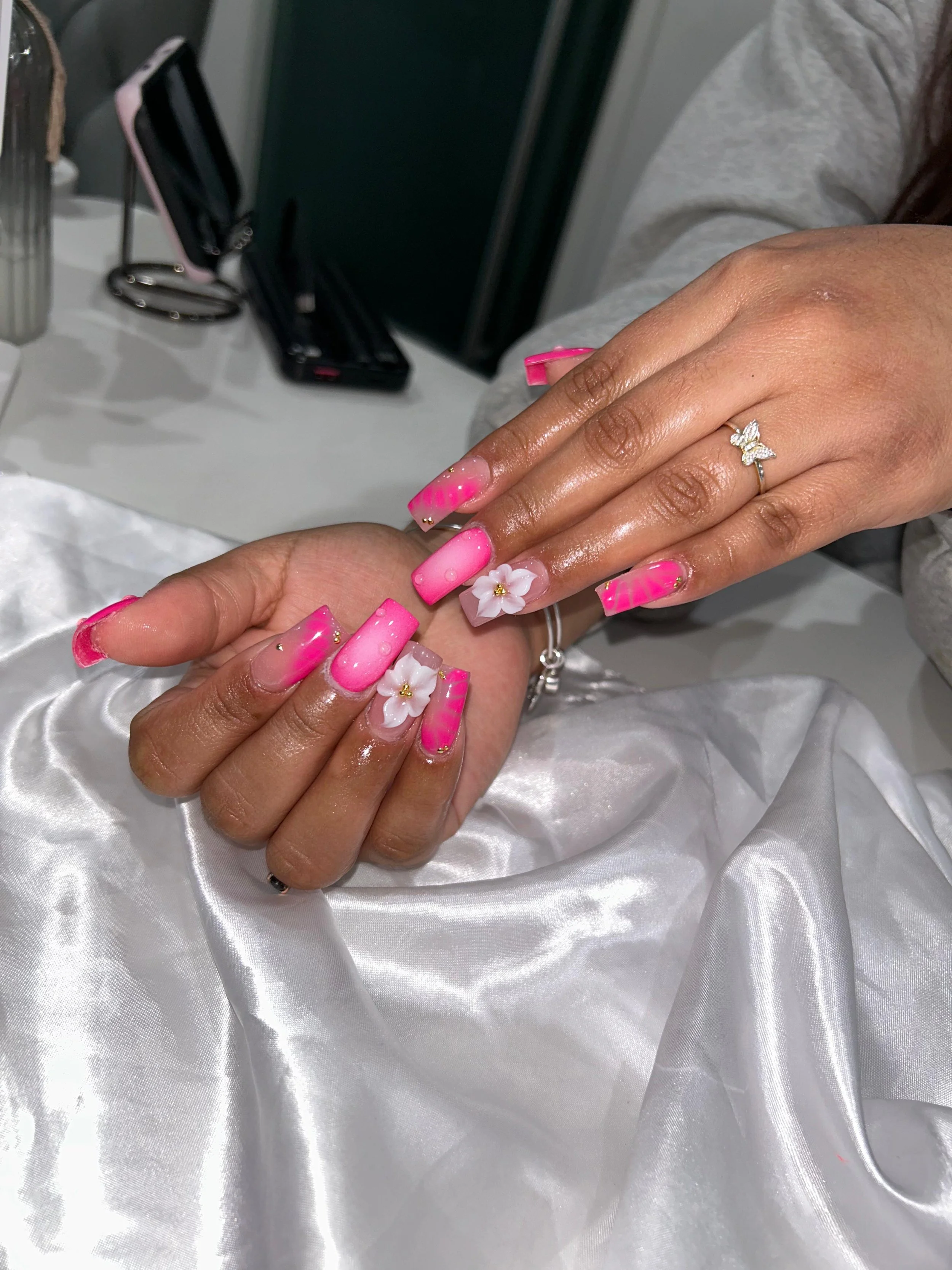 Close-up of a woman with pink and white nail art, with 3D flower embellishments, showing a ring with a butterfly design on her finger, and another hand with similar nail art resting on a silky white cloth.