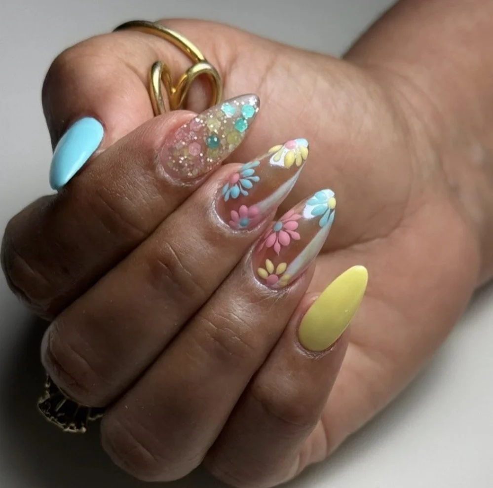 Colorful manicured nails with floral and glitter designs, with a gold ring on the finger.