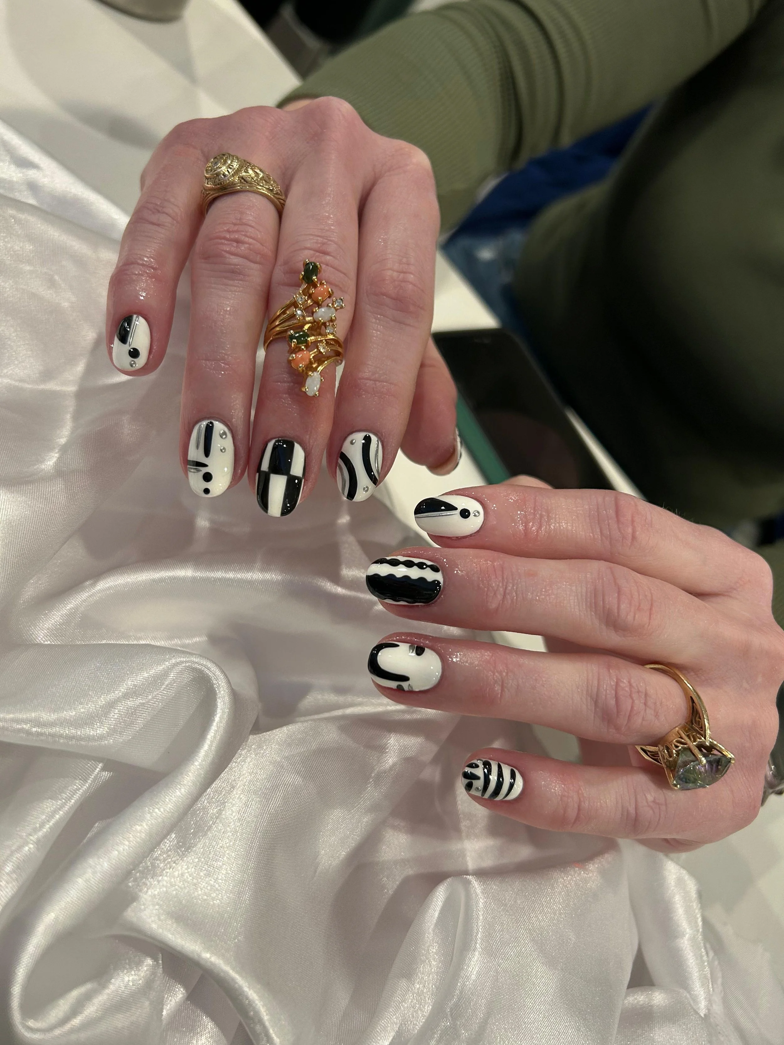 Close-up of a person's hands with black and white abstract patterned nail art and decorated with multiple gold rings, resting on a satin fabric.