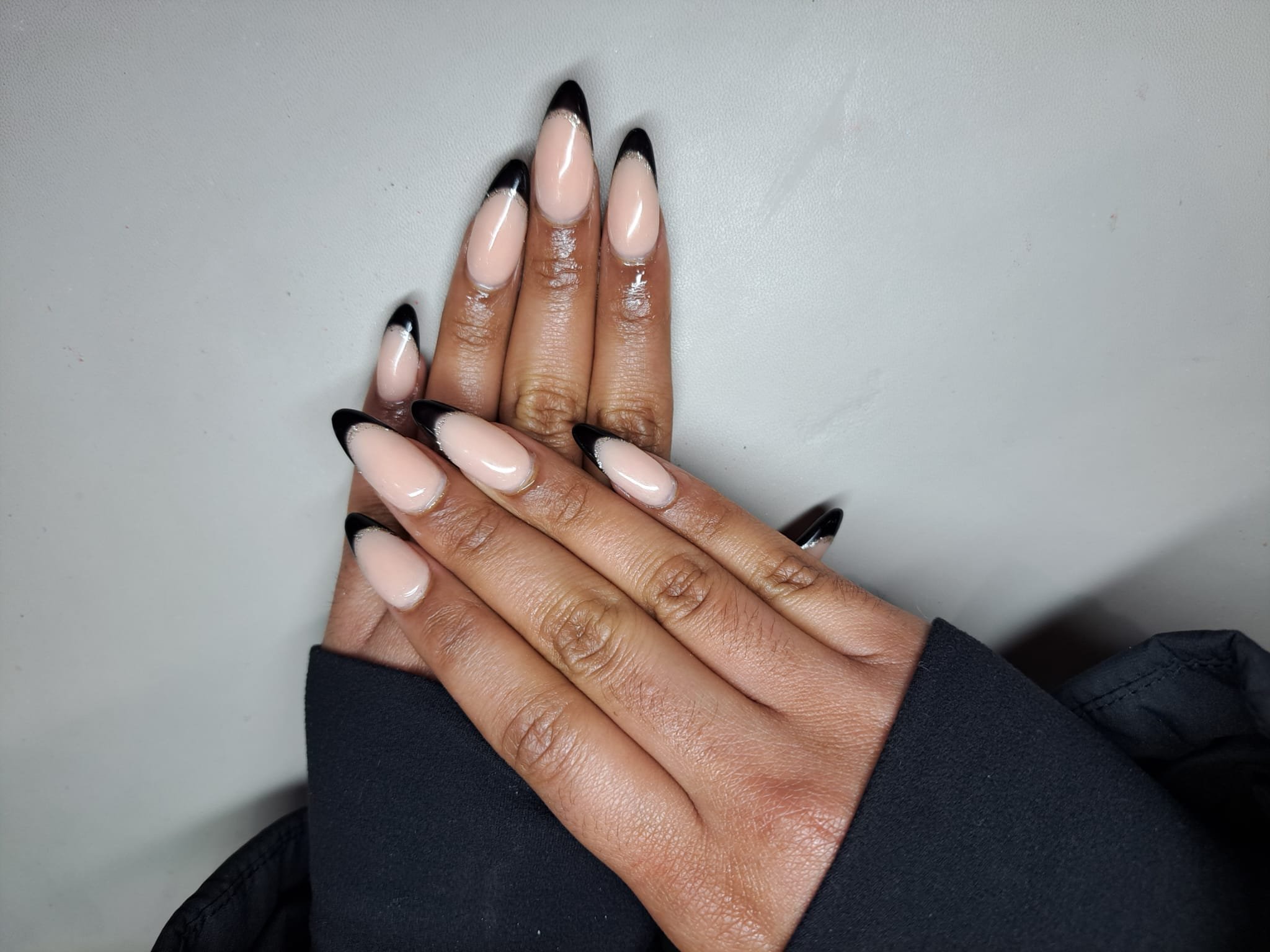 A person's hands with manicured nails featuring a nude base color and black tips, resting on a gray surface.