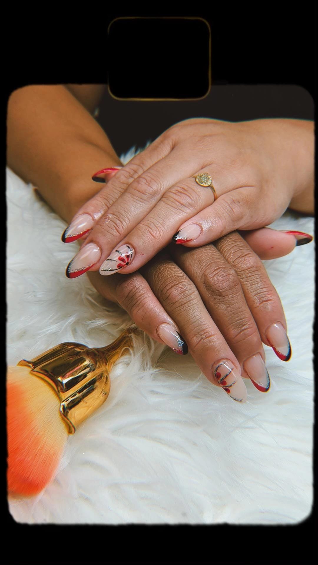 Close-up of two hands with manicured nails featuring red, black, and glittery tips, resting on a white furry surface. One hand has a gold ring with a stone. A makeup brush with orange powder is in the foreground.
