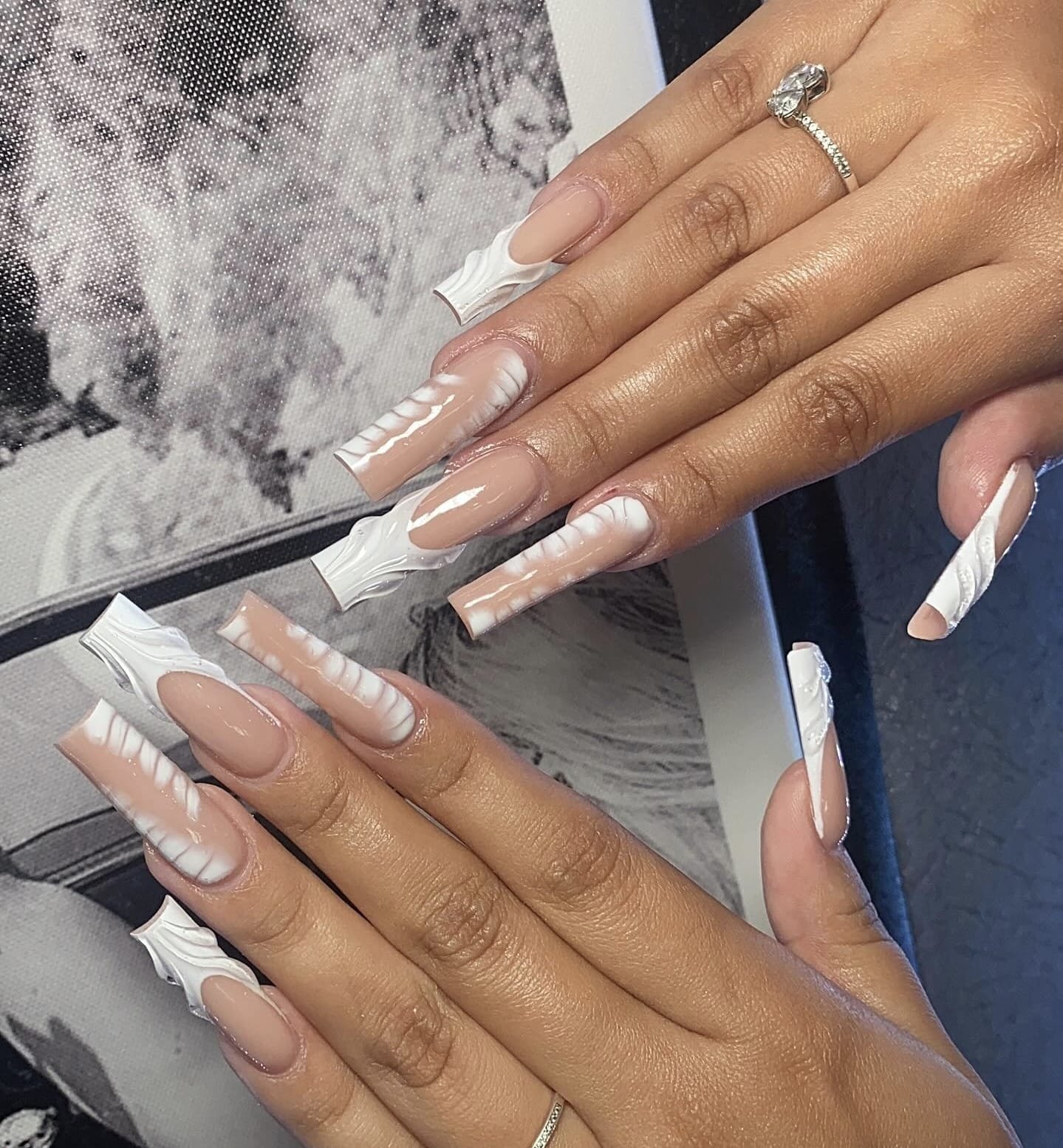 Close-up of hands with long acrylic nails featuring a swirl design in white and nude colors, and a ring on the middle finger of the right hand.