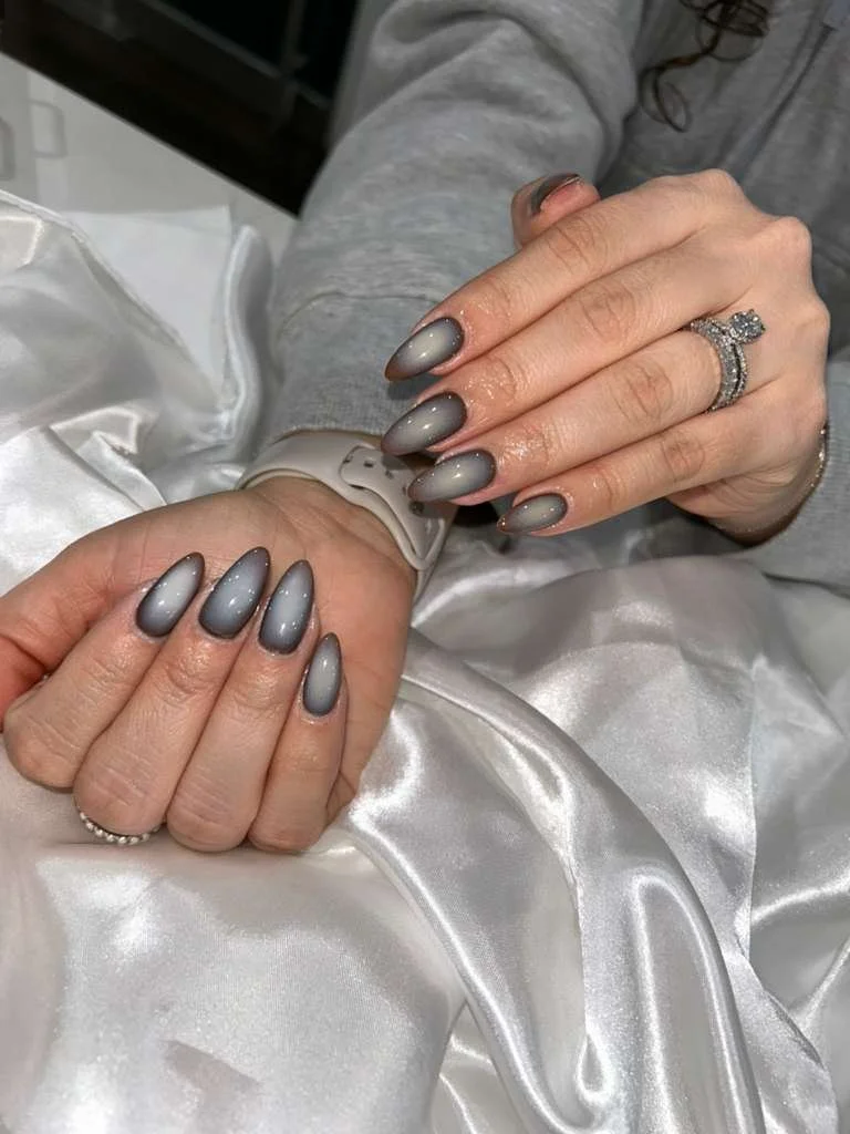 Person showing manicured nails with gray to black gradient polish, wearing a silver ring with a large gemstone, and a white watch, resting on shiny white satin fabric.