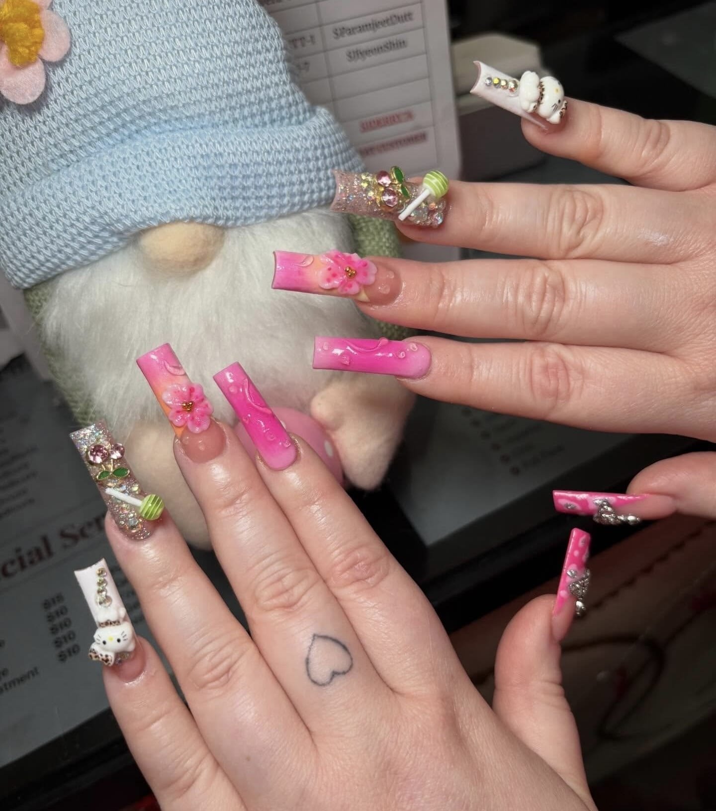 Close-up of hands with elaborate pink and white 3D nail art, decorated with flowers, rhinestones, and charms, in front of a plush gnome toy.