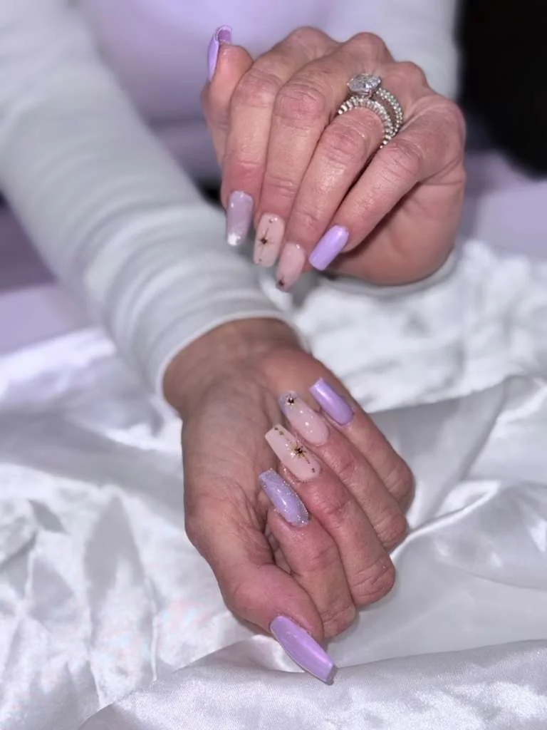 Close-up of two hands with manicured nails, adorned with rings, resting on a silky white fabric. Nails are painted in pastel colors with some featuring small decorative elements.