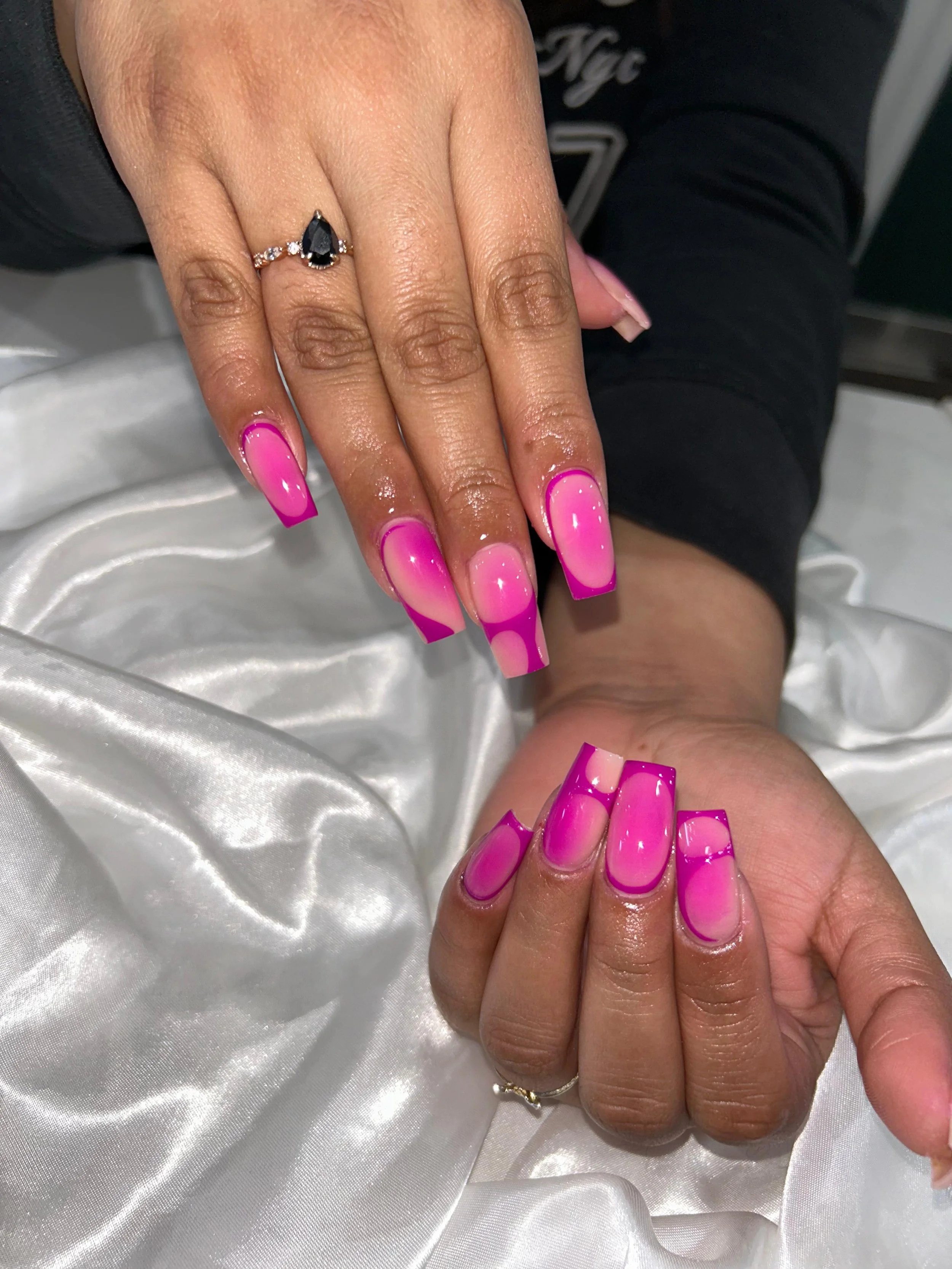 Close-up of a person's hands and nails with bright pink and purple nail polish and a black teardrop-shaped ring with small diamonds on the left hand.