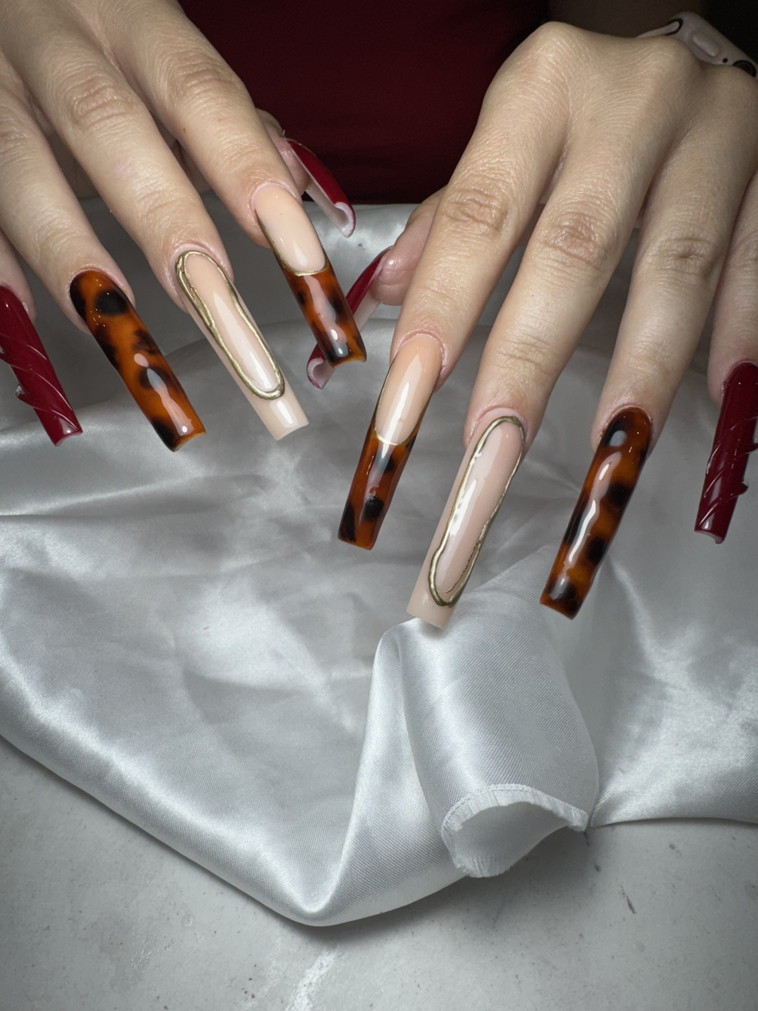 Close-up of hands with long, square-shaped acrylic nails featuring tortoiseshell, nude, and red polish designs with gold accents, resting on a satin fabric background.