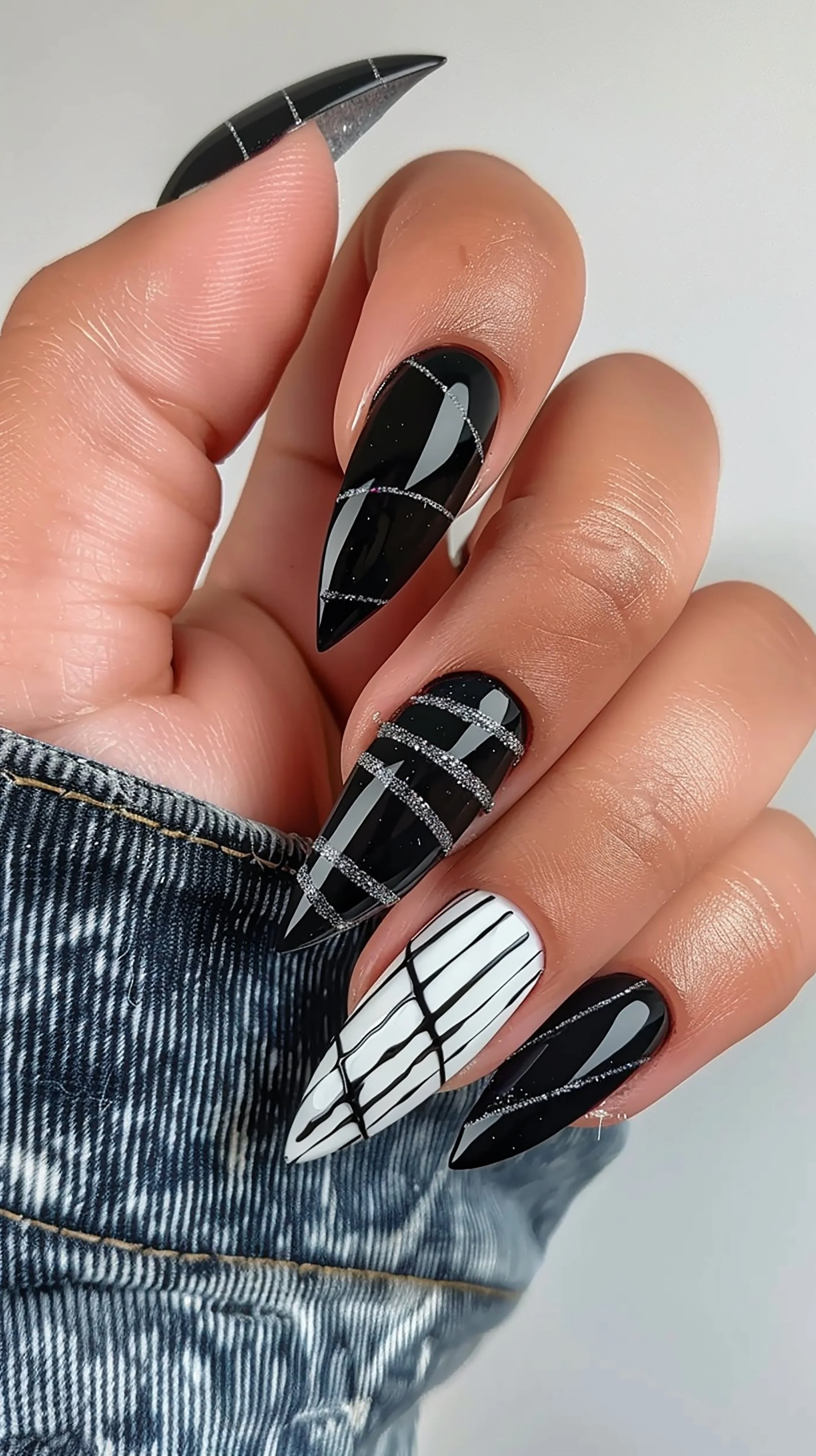 Close-up of a hand with long, pointed black and white designer acrylic nails with plaid and stripe patterns, also featuring silver glitter accents, wearing a denim jacket.
