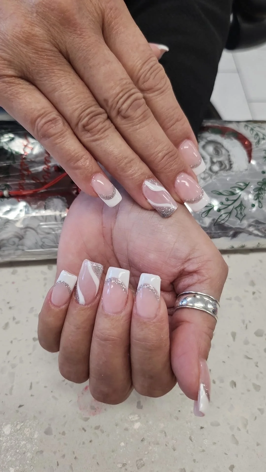 Hands with decorated French manicure nails, featuring silver glitter and white swirling design, resting on a speckled countertop with a Christmas-themed gift bag in the background.