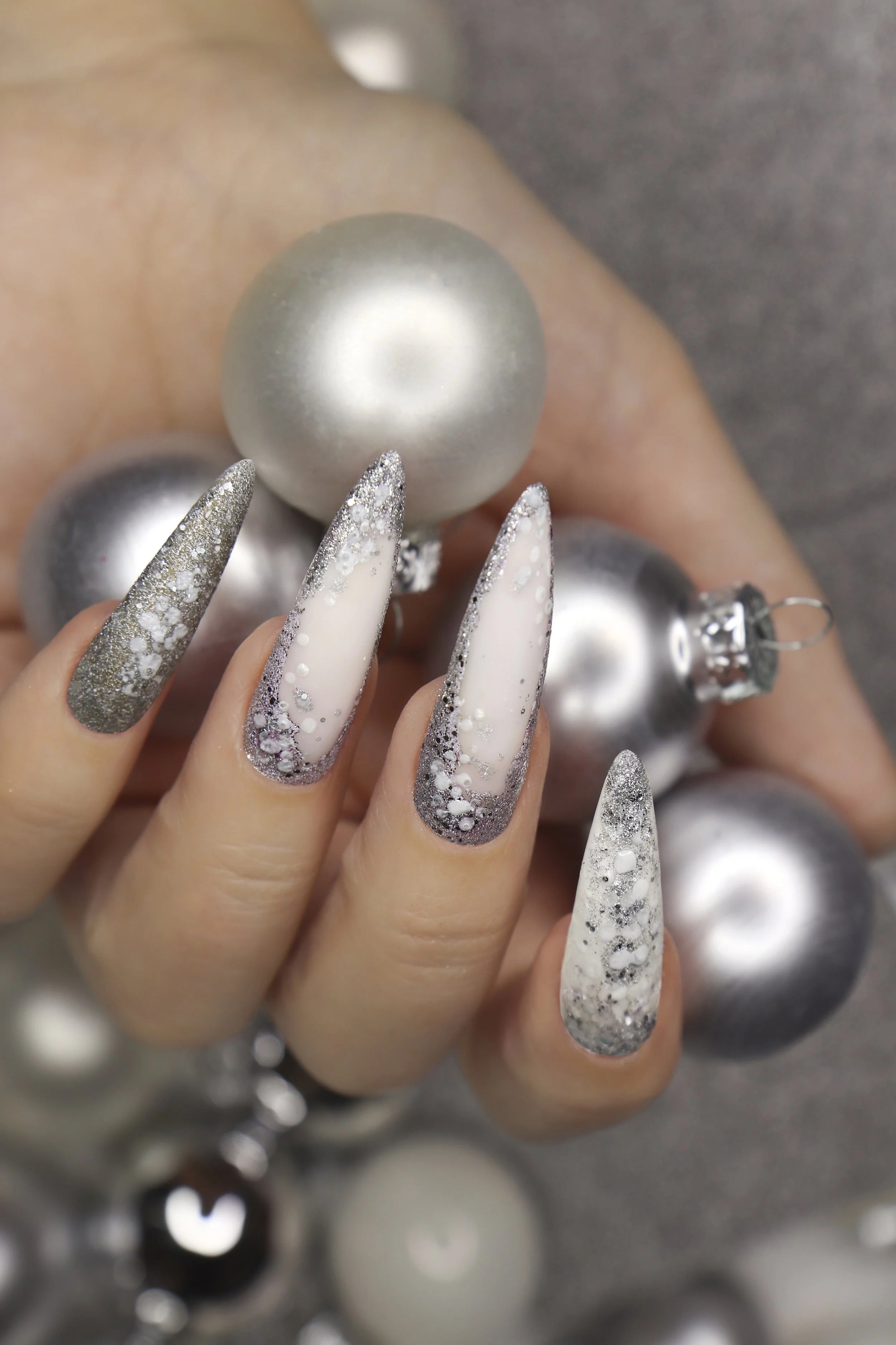 Close-up of a hand with long, almond-shaped, decorated nails in silver, white, and black, holding several shiny Christmas ornaments, including silver and white baubles, in the background.