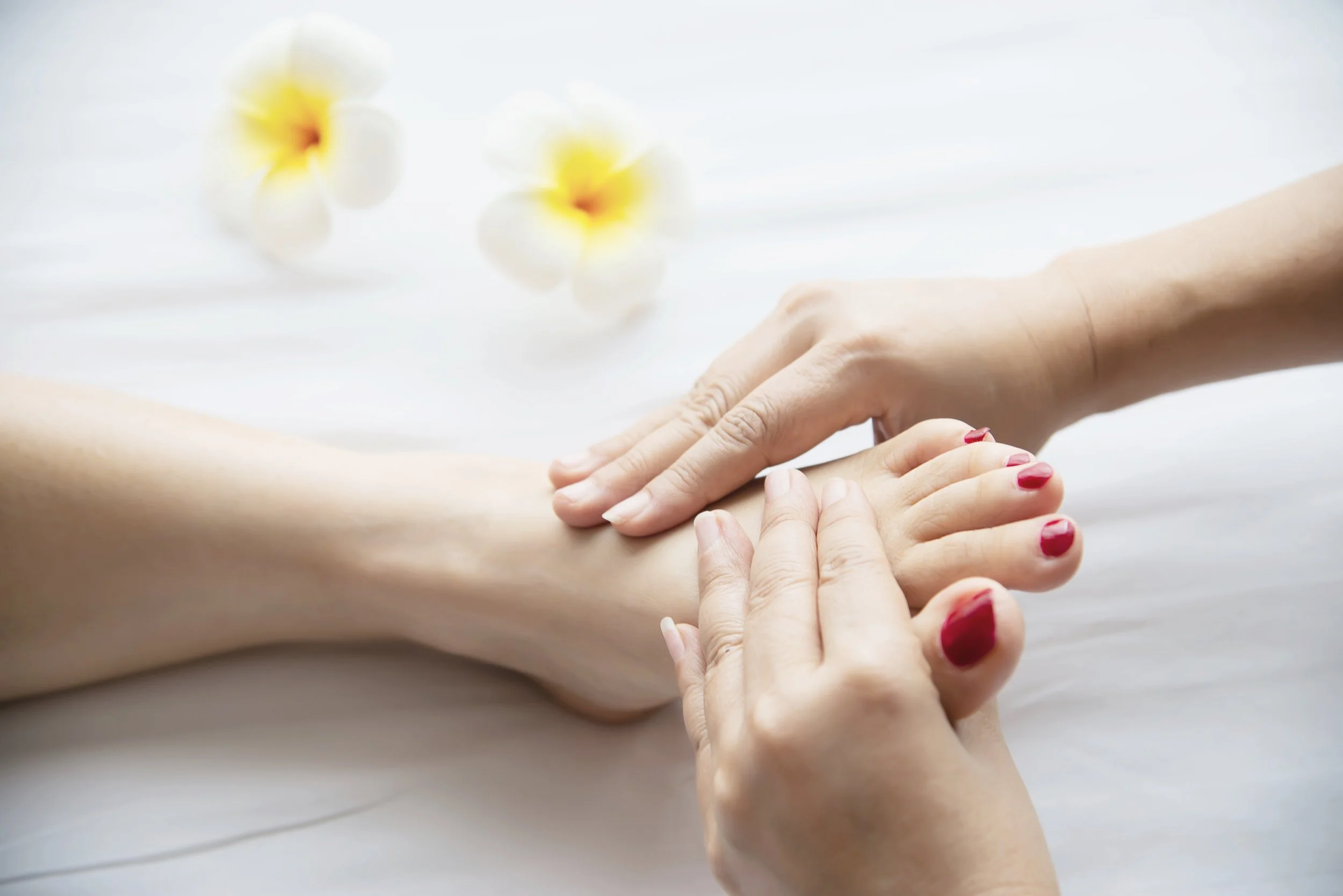 A person receiving a massage, with hands on their forearm and hand, on a white surface with white and yellow flowers in the background.