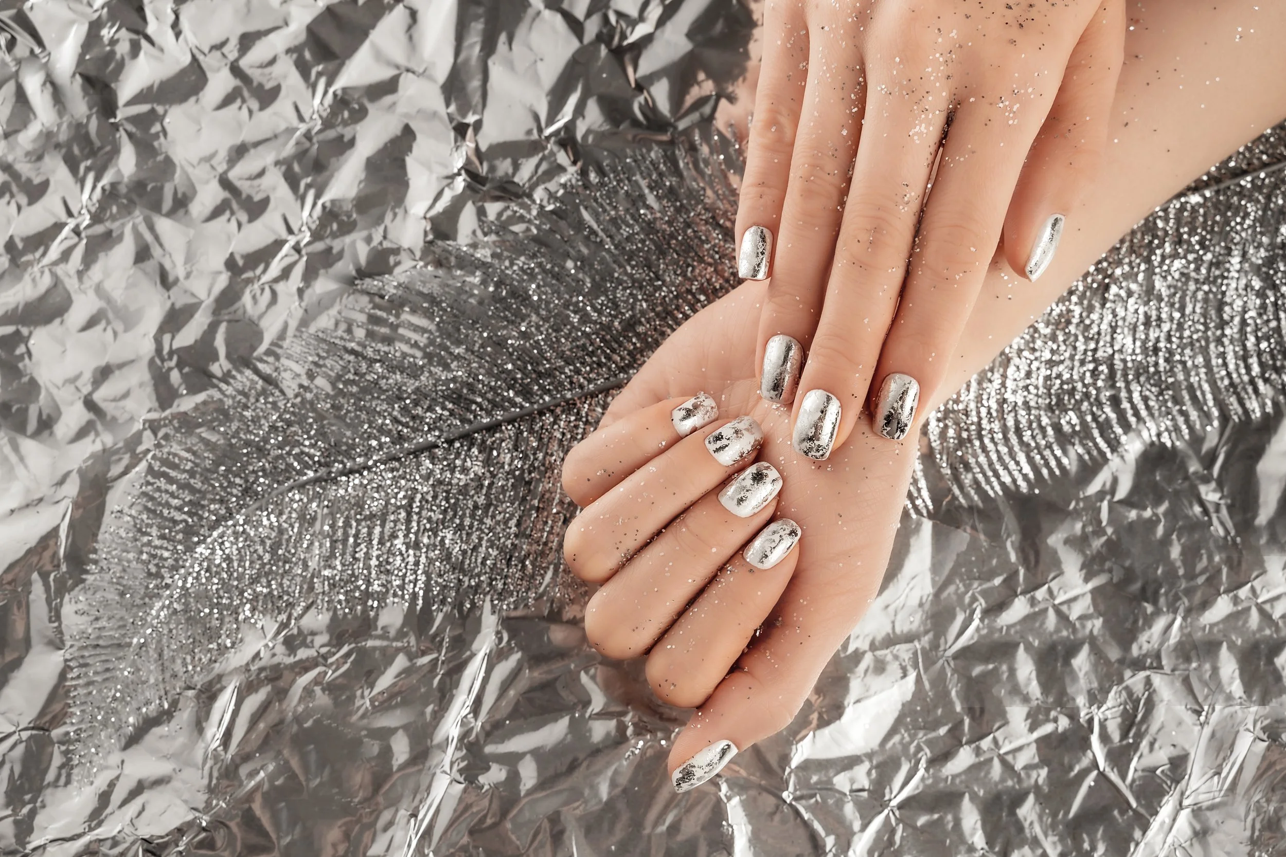 Hands with metallic silver nail polish and black marble design, resting on crumpled metallic silver foil with a large reflective silver feather.