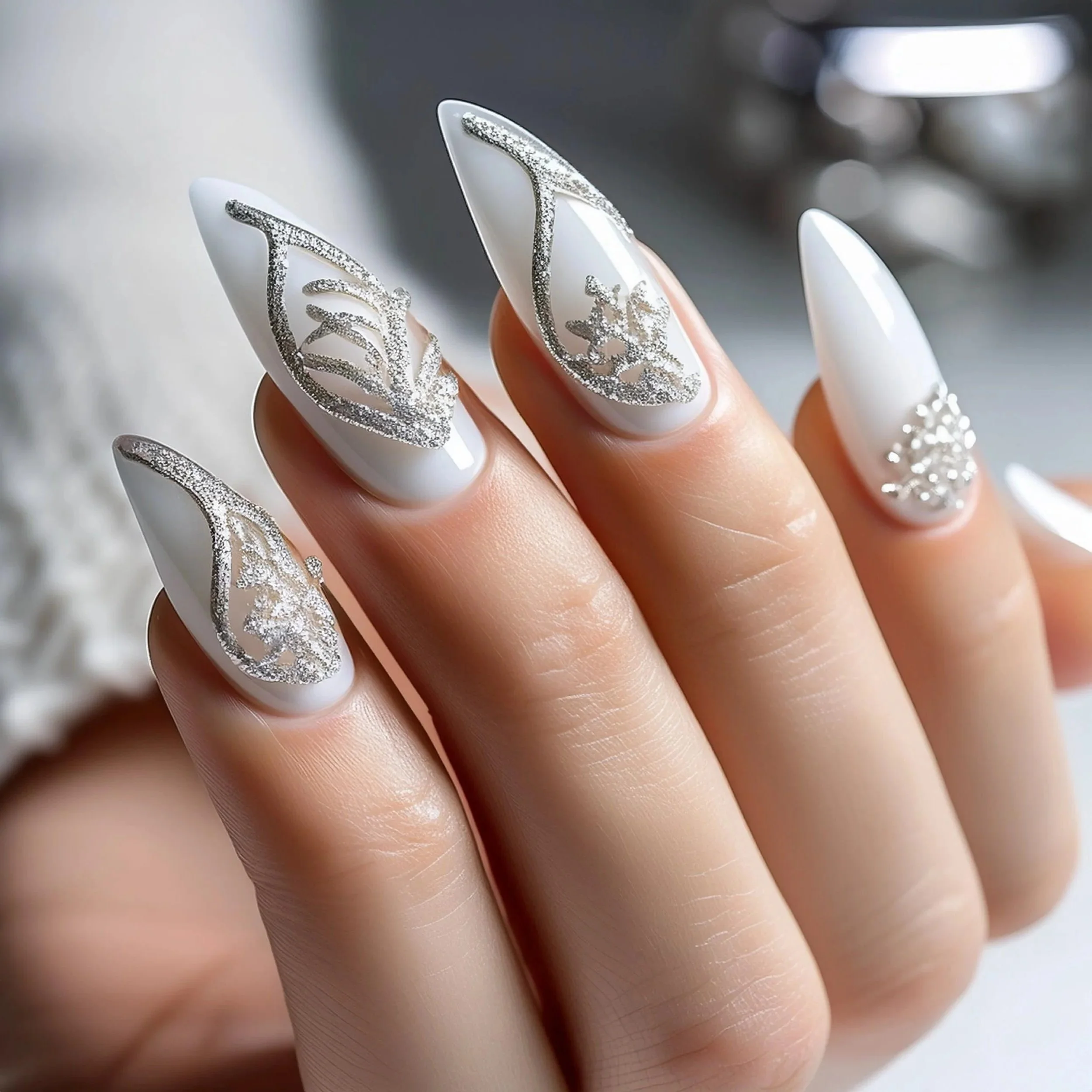 Close-up of a hand with white, almond-shaped nails decorated with silver glittery intricate designs.