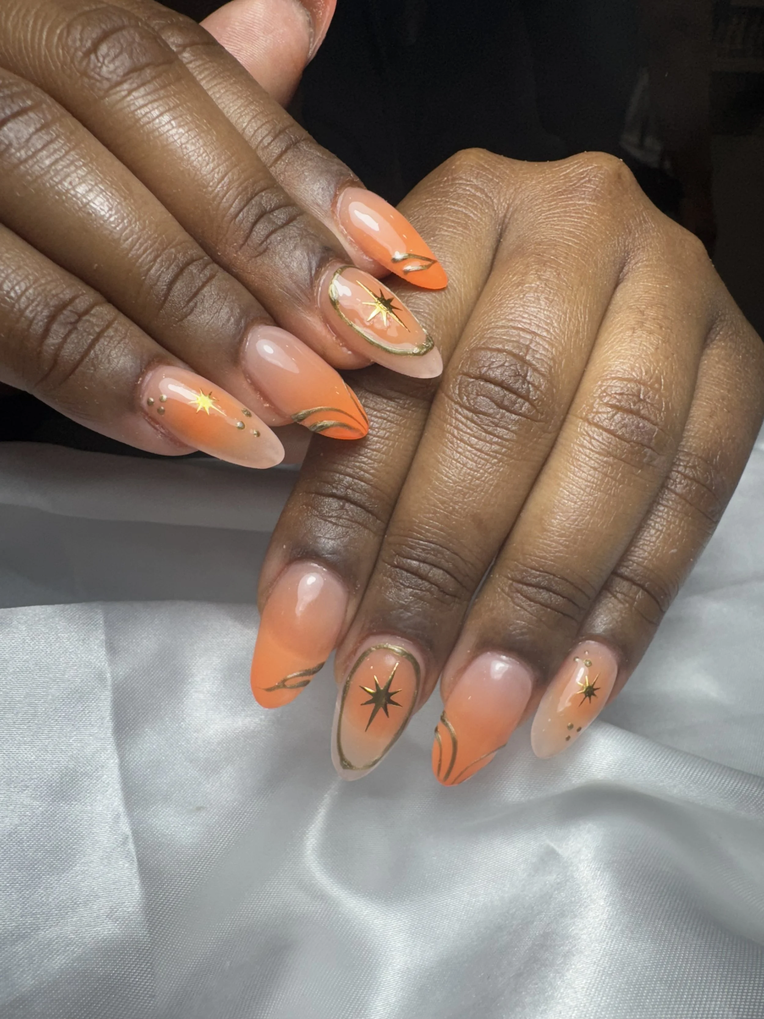 Close-up of hands with orange and nude ombre nails decorated with gold star and swirl designs.