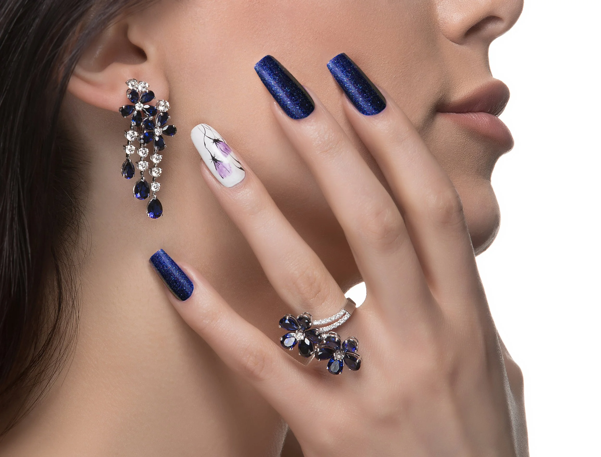 Close-up of a woman's face with a focus on her jewelry, including a floral ring, an earring with black and clear gemstones, and a ring with black and clear stones, and her nails painted dark blue with a glittery finish. She has a white floral nail art on her middle finger.