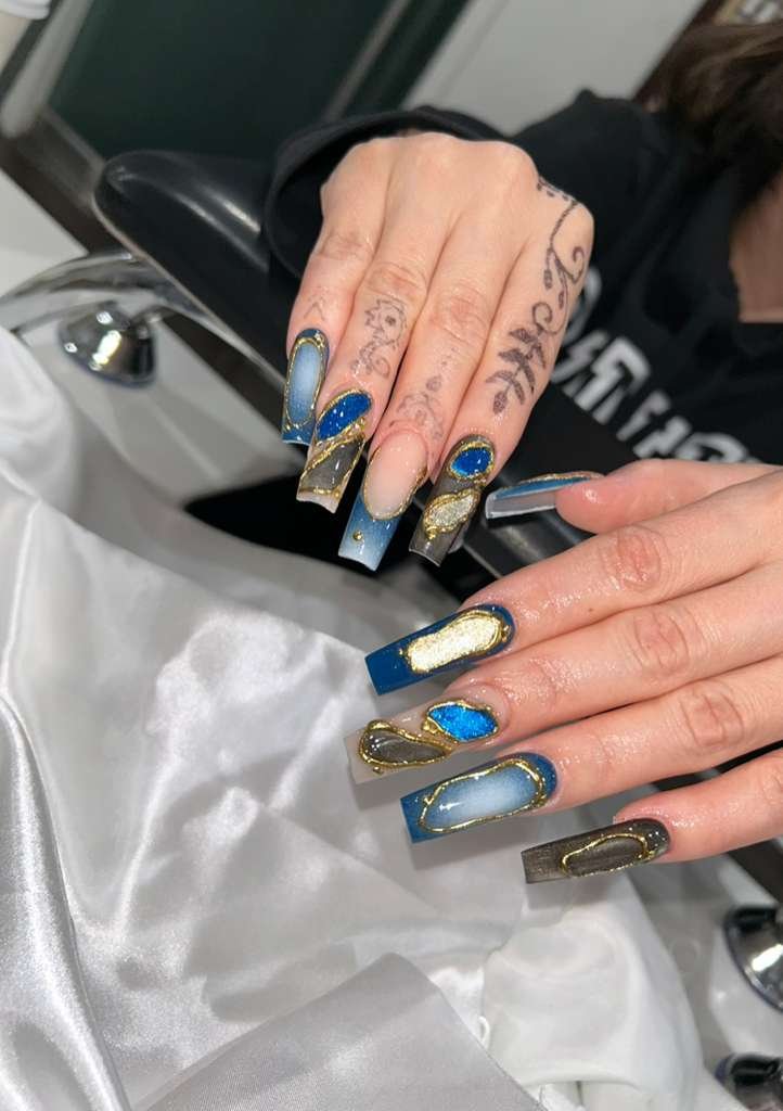 Close-up of nails with elaborate blue, gold, and silver design, on a person's hand resting on a white cloth at a salon.